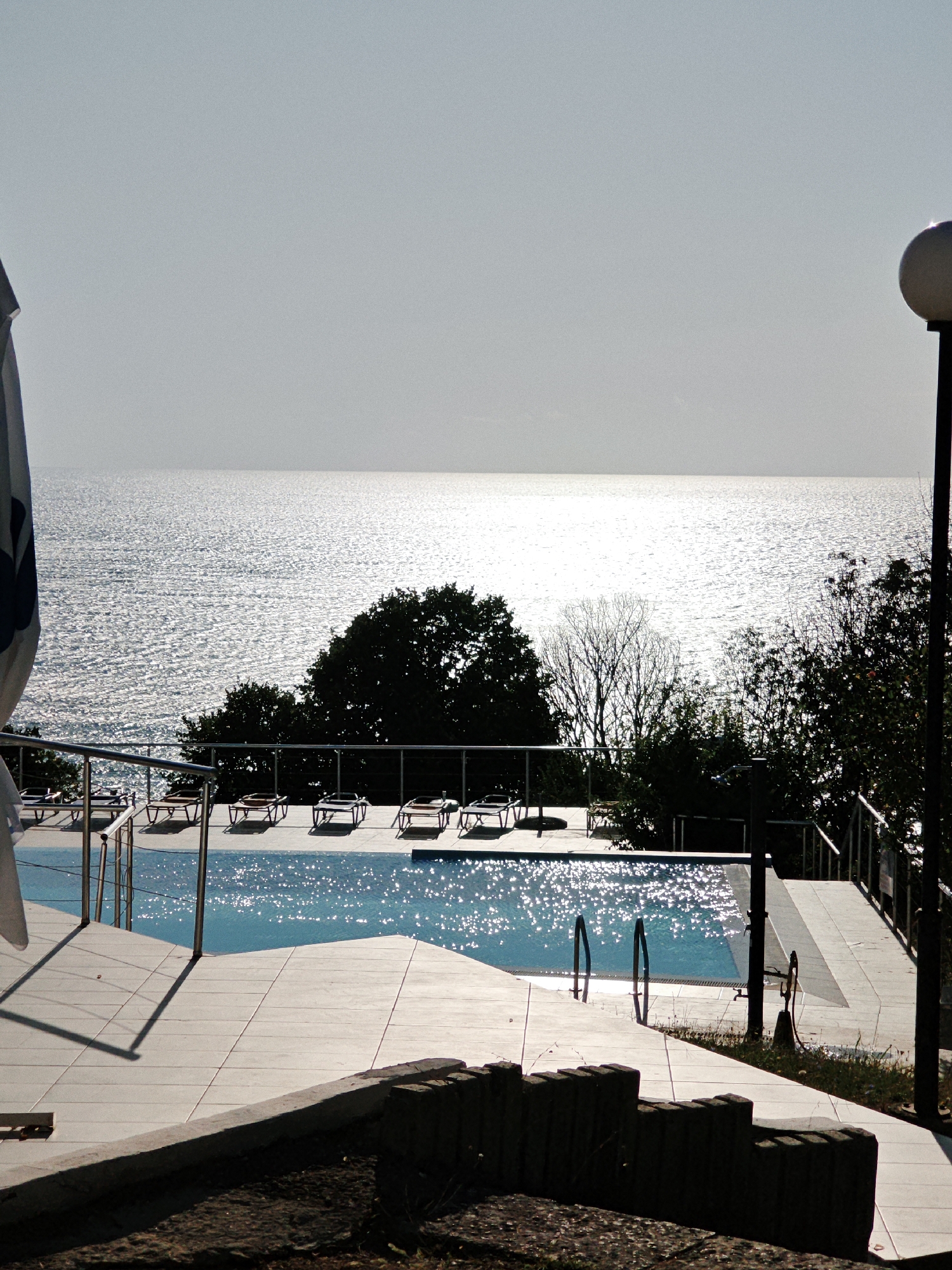 This is a photograph of a swimming pool area overlooking a body of water. The pool is rectangular and filled with blue water that reflects the sunlight. There are several lounge chairs lined up around the pool. The area surrounding the pool is paved with light-colored tiles. In the background, there is a dense line of trees, and beyond that, the expanse of the ocean or a large lake. The sky is a pale blue, almost white, indicating bright daylight conditions. A streetlamp is visible to the right of the frame. The foreground shows a rough, uneven surface, possibly a garden or a pathway. The overall scene suggests a resort or a private residence with a view of the water.