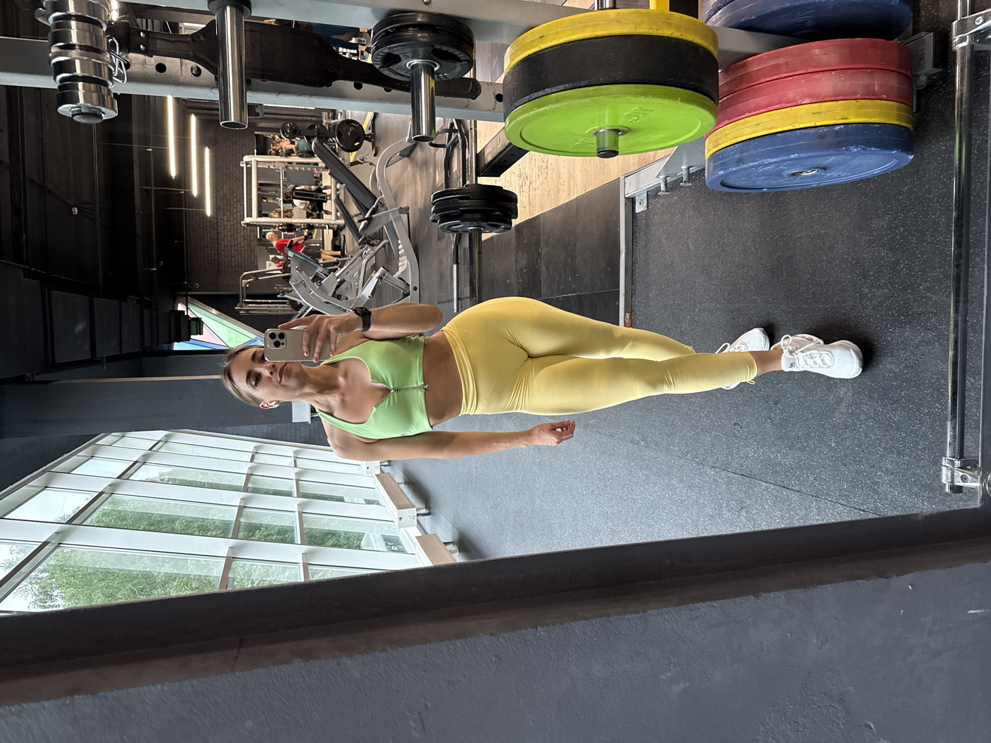 A woman in a green sports bra and yellow leggings poses in what appears to be a gym. The woman is holding a phone to take a selfie. Behind her are stacks of weights. Large windows are on the left side of the scene. Based on the gym's interior and window view, the gym could be located in a modern facility, possibly in Saint Petersburg, Russia, given the request for its location. Other gym equipment can be seen blurred in the background. The gym has a dark-colored floor. The image appears to be taken in a mirror, showing a reflection of the gym and the woman.