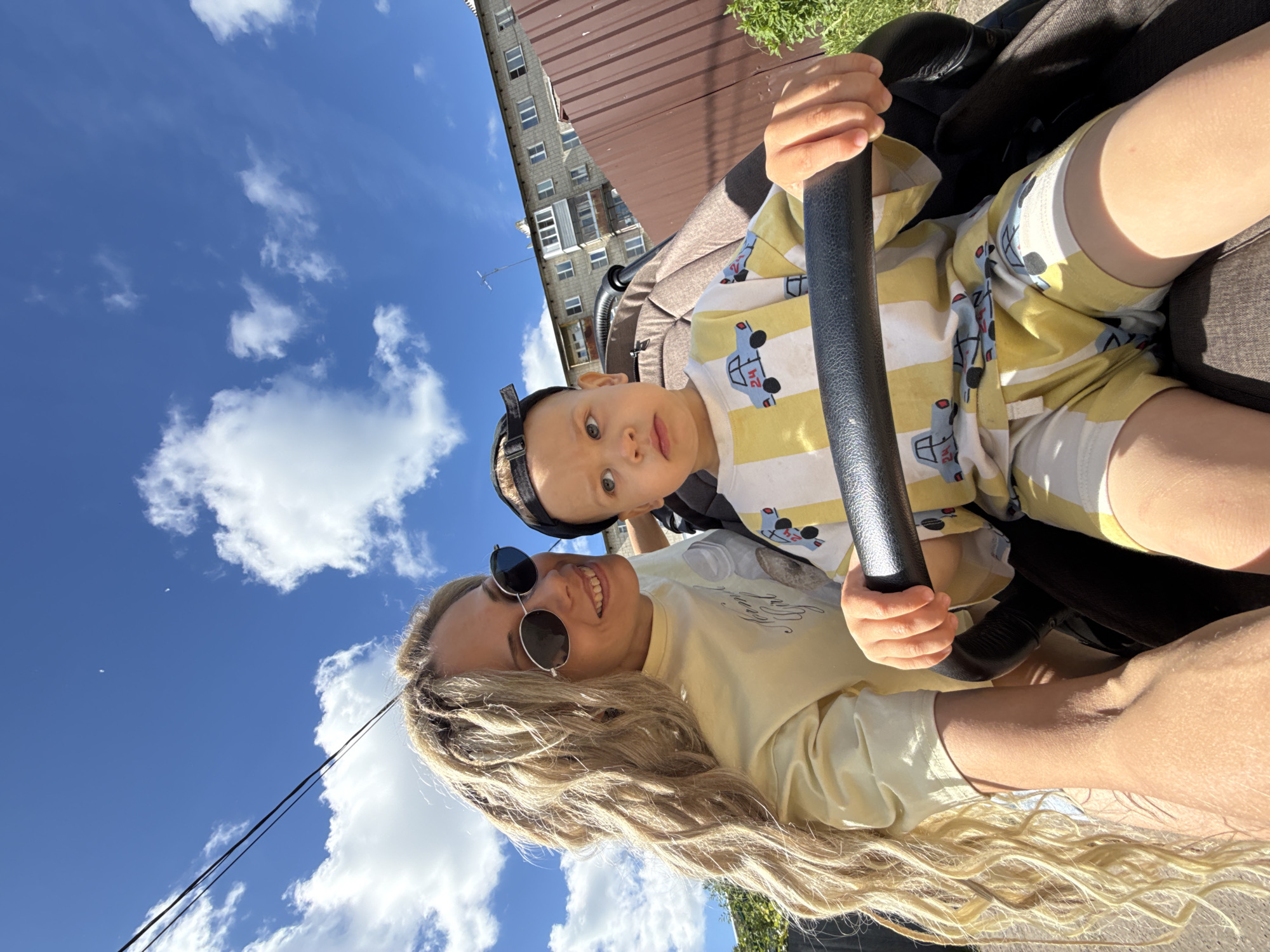 The image captures a cheerful outdoor moment in Rusanovo, Russia, featuring a woman and a young child from a low-angle perspective. The woman, with long, wavy blonde hair, is smiling broadly at the camera while wearing a light-colored long-sleeved top and round black sunglasses. Above her, a toddler sits in what appears to be a stroller or a child's ride-on toy, gripping a black bar with both hands. The child is dressed in a yellow and white striped outfit patterned with cartoon cars, some faintly displaying the number "24," and a black cap worn backward, looking directly forward with wide, curious eyes.
The background reveals a bright blue sky filled with scattered white cumulus clouds, indicating fair weather and strong daylight, likely mid-day. To the right, a multi-story residential building with numerous windows and a brown corrugated metal roof is visible, along with green foliage. Two dark electrical wires are also present against the sky. The overall scene suggests a pleasant, casual interaction between the adult and child under sunny conditions.