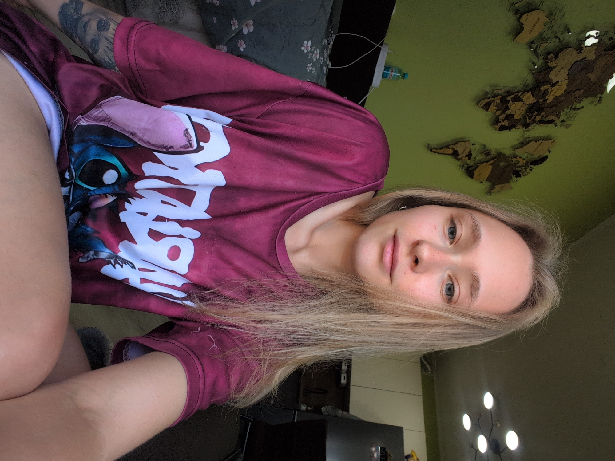A young woman with blonde hair is seated indoors, facing the camera. She is wearing a burgundy t-shirt with a graphic print of Stitch from Lilo & Stitch and white lettering. The woman has tattoos on her left arm, including one of Groot from Guardians of the Galaxy. She is positioned in front of a green wall adorned with a wooden world map. To the left of the woman, a bed with a floral-patterned duvet is visible. In the background, a kitchen area can be seen, with cabinets, a refrigerator, and a sink. A ceiling light fixture with multiple bulbs is also present. The lighting suggests it is daytime, with natural light entering the room.