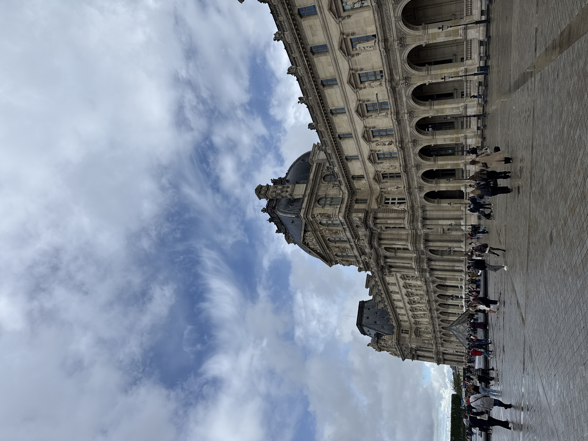 The image shows a view of the Louvre Museum in Paris, France. The building is a large, ornate structure with multiple arched windows and detailed architectural features. A crowd of people is gathered in front of the building, likely tourists or visitors. The sky is overcast with dramatic cloud formations. The foreground consists of paved ground reflecting light, possibly wet from recent rain.