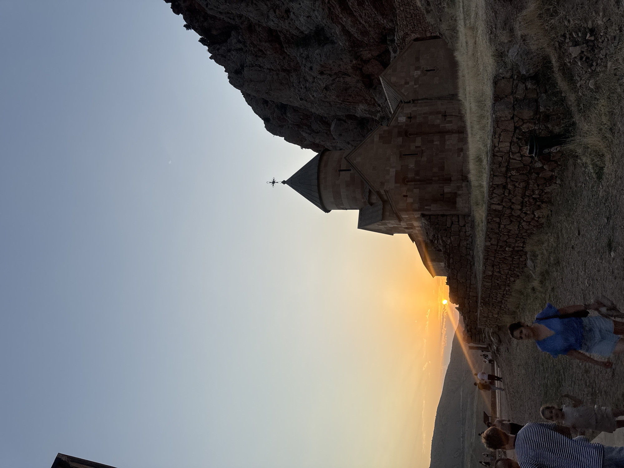An outdoor, low-angle, vertical shot shows the Noravank monastery in Armenia at sunset. The sun is setting behind a hill in the background, casting a warm glow over the scene. The monastery, made of reddish-brown stone, is the main subject, with its conical roof and cross prominently displayed. A few people are visible in the foreground, walking on a dirt path. The monastery is situated in a rocky, mountainous landscape.