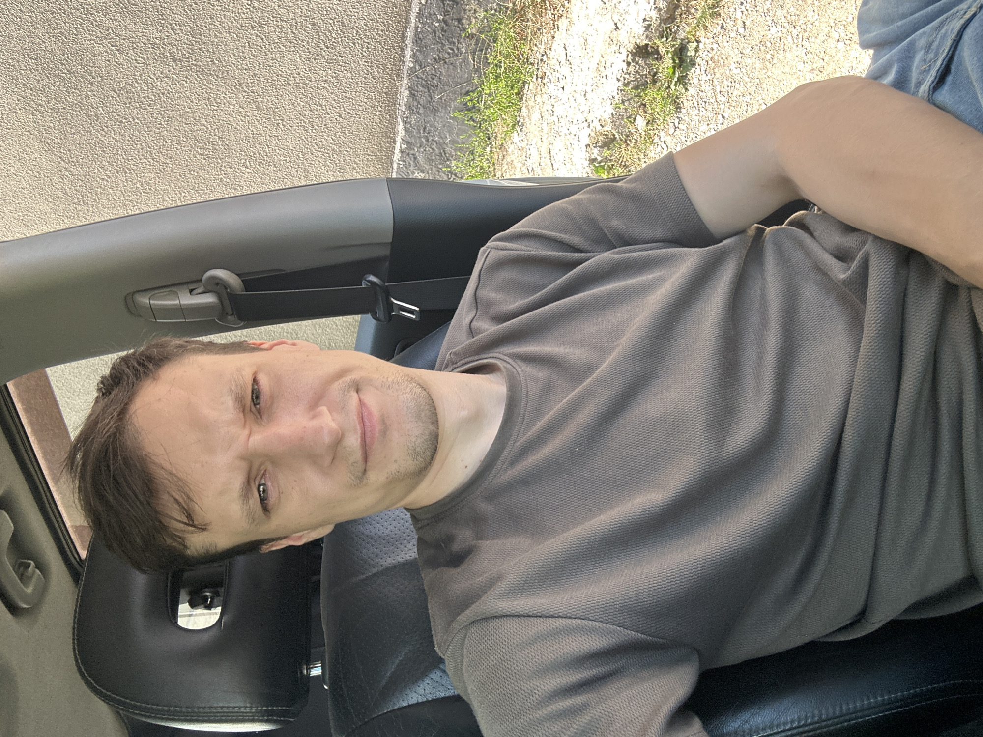 A man with short brown hair and light skin is sitting in the driver's seat of a car, looking at the camera. He is wearing a grey t-shirt and blue jeans. The interior of the car is visible, including a black leather seat and a black headrest. Outside the car, a textured tan wall and some grass and gravel are visible. The man's facial expression is neutral, with slightly furrowed brows. The overall lighting suggests it is daytime.