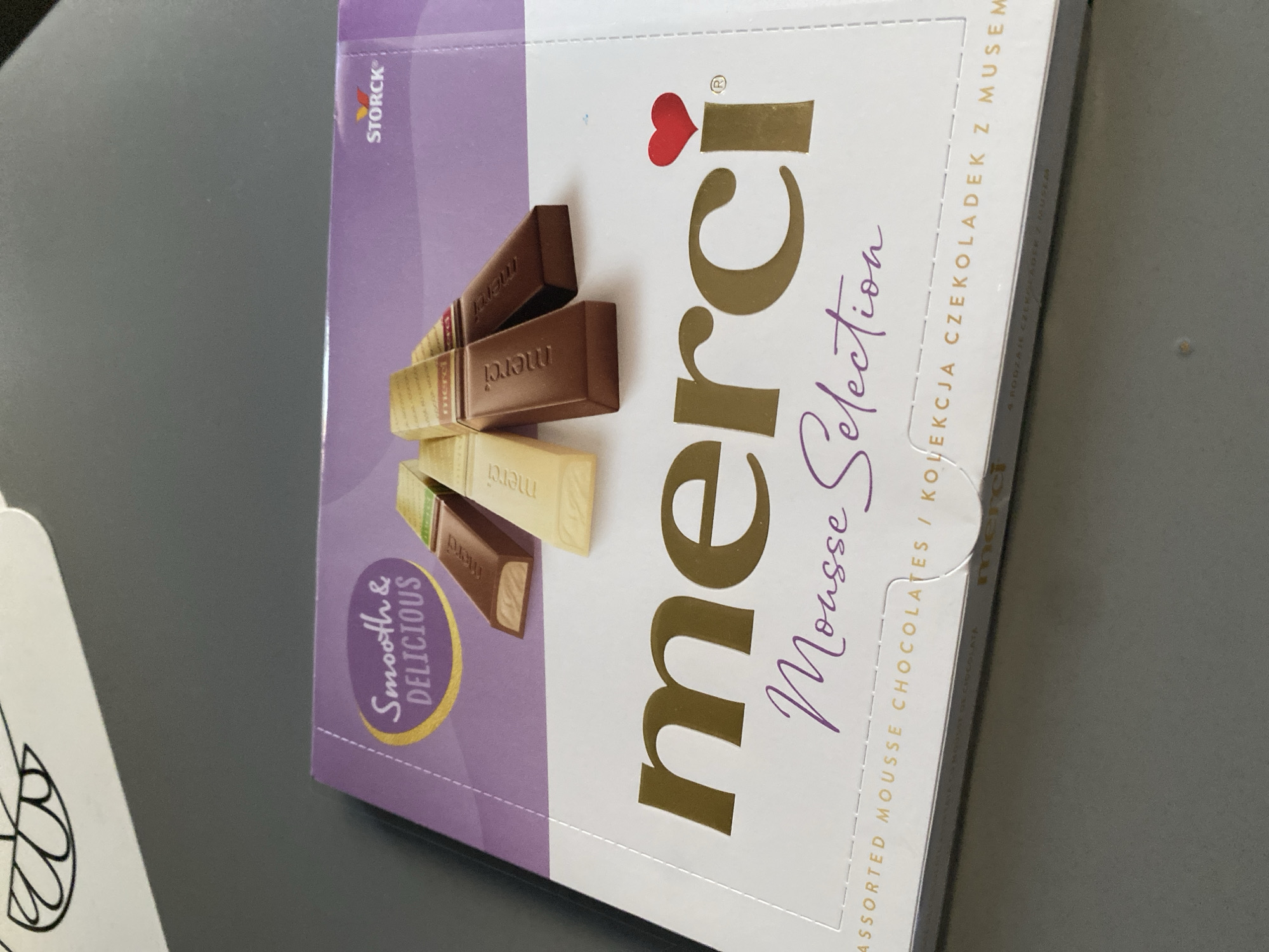 The image shows a close-up of a box of Merci Mousse Selection chocolates. The box has a purple and white design with the Merci logo and the words "Smooth & Delicious" prominently displayed. Several individual chocolate bars are visible, including milk chocolate and white chocolate varieties. The box is resting on a gray surface, possibly a table or countertop. The background is out of focus. There is no indication of the city of Wrocław, Poland, or any other specific location.