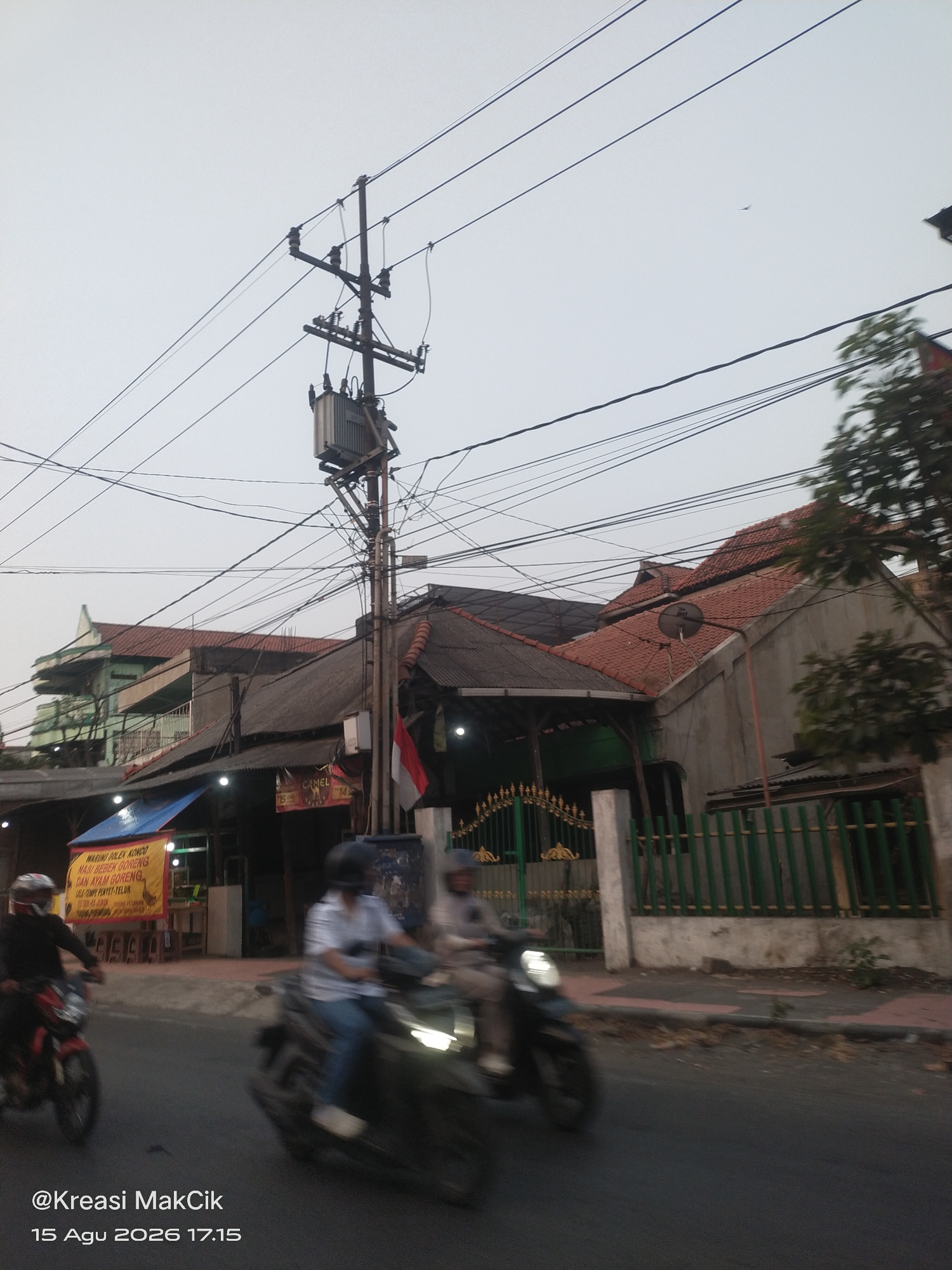 The image depicts a street scene in Wonocolo, Indonesia, possibly on August 15, 2026, at 17:15. In the foreground, two motorcycles are in motion, their riders blurred due to the movement. The motorcycle on the left is red and black, with a rider wearing a helmet and dark clothing. The motorcycle on the right is dark colored, with a rider in a light-colored shirt and blue pants, also wearing a helmet.
The background reveals a row of buildings with terracotta tiled roofs. One building has a prominent green facade and multiple levels. Another building features a corrugated metal roof and an attached Indonesian flag. In front of some of these buildings are open-air stalls, one displaying a yellow banner with red text. Electrical poles with a dense network of wires and a transformer are visible above the buildings, indicating urban infrastructure. A green metal fence with decorative elements runs along the sidewalk in front of some of the properties. The sky is overcast and light-colored. Text overlays at the bottom of the image read "@Kreasi MakCik" and "15 Agu 2026 17.15".