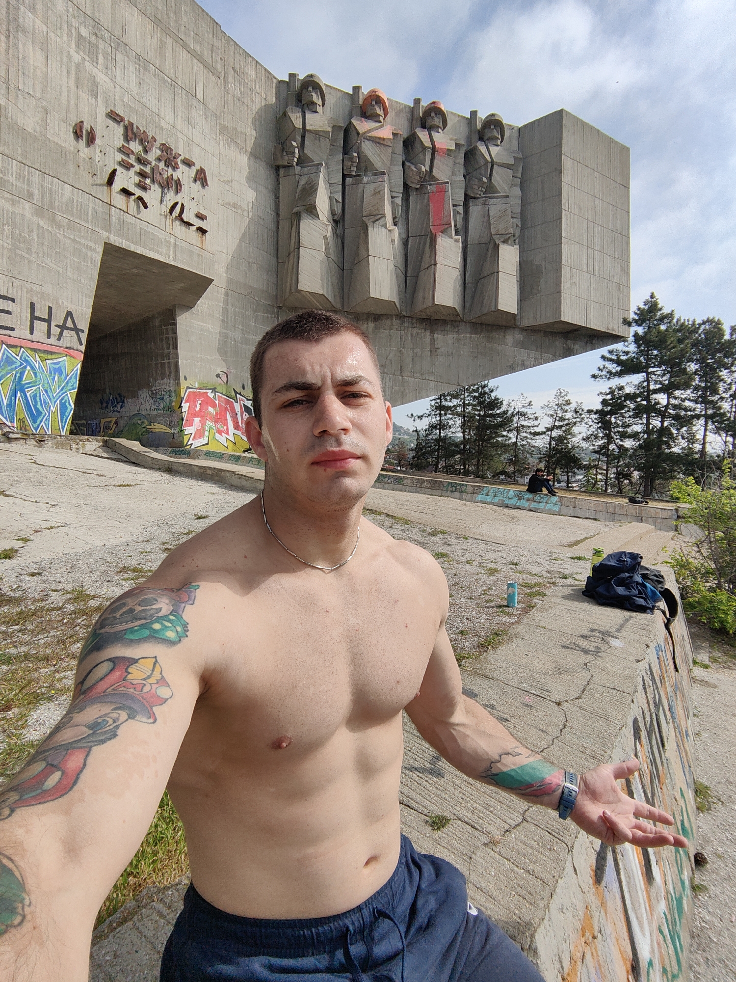 A shirtless man stands in front of the Monument of the Bulgarian-Soviet Friendship in Varna, Bulgaria, taking a selfie. He has short dark hair, a silver necklace, and tattoos on both arms. The monument is a large concrete structure with four figures at the top. There is graffiti on the monument and the surrounding area. In the background, there are trees and people sitting on a ledge. A water bottle and a bag are next to the ledge. The sky is partly cloudy.