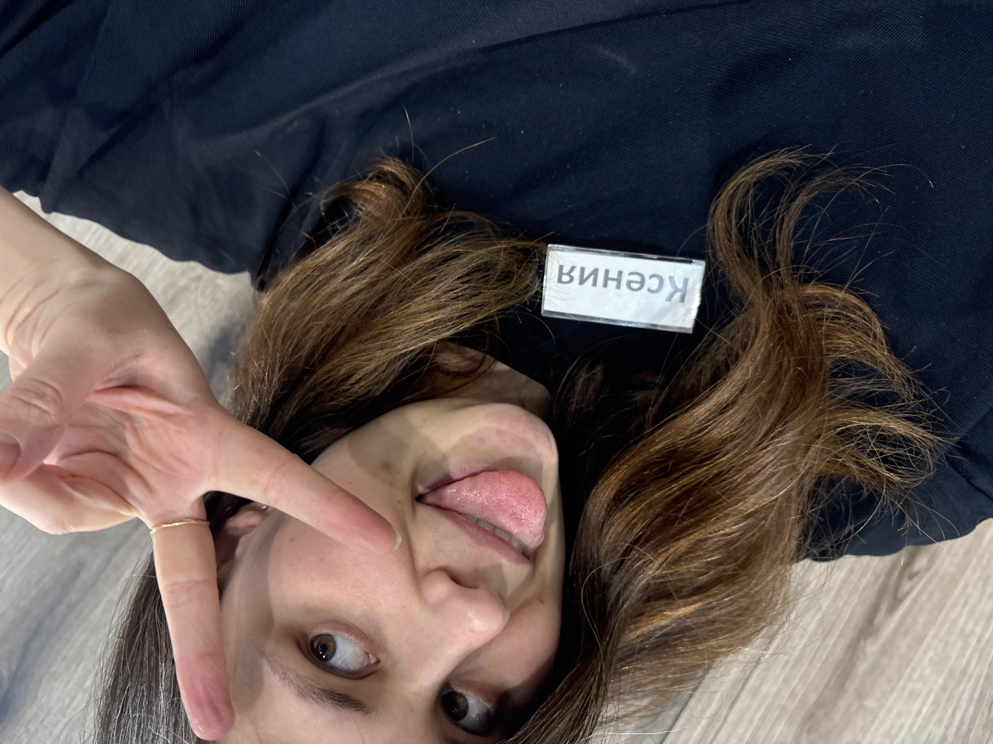 A forensic examination of the image reveals a female individual identified as Ksenia, located in Chaikovsky, Russia. The subject is depicted in an indoor setting, observed from a high-angle perspective, against a light wood-effect plank floor. She possesses brown hair and brown eyes, and is wearing a black short-sleeved top. Her right hand is raised to eye level, forming a "V" sign with her index and middle fingers. A thin, gold-colored ring is visible on the ring finger of her right hand. Her tongue is extended from her mouth, creating a playful expression. A rectangular name tag, clipped to the left side of her black top, displays the Cyrillic name "Ксения" (Ksenia). The subject's gaze is directed upwards within the frame.