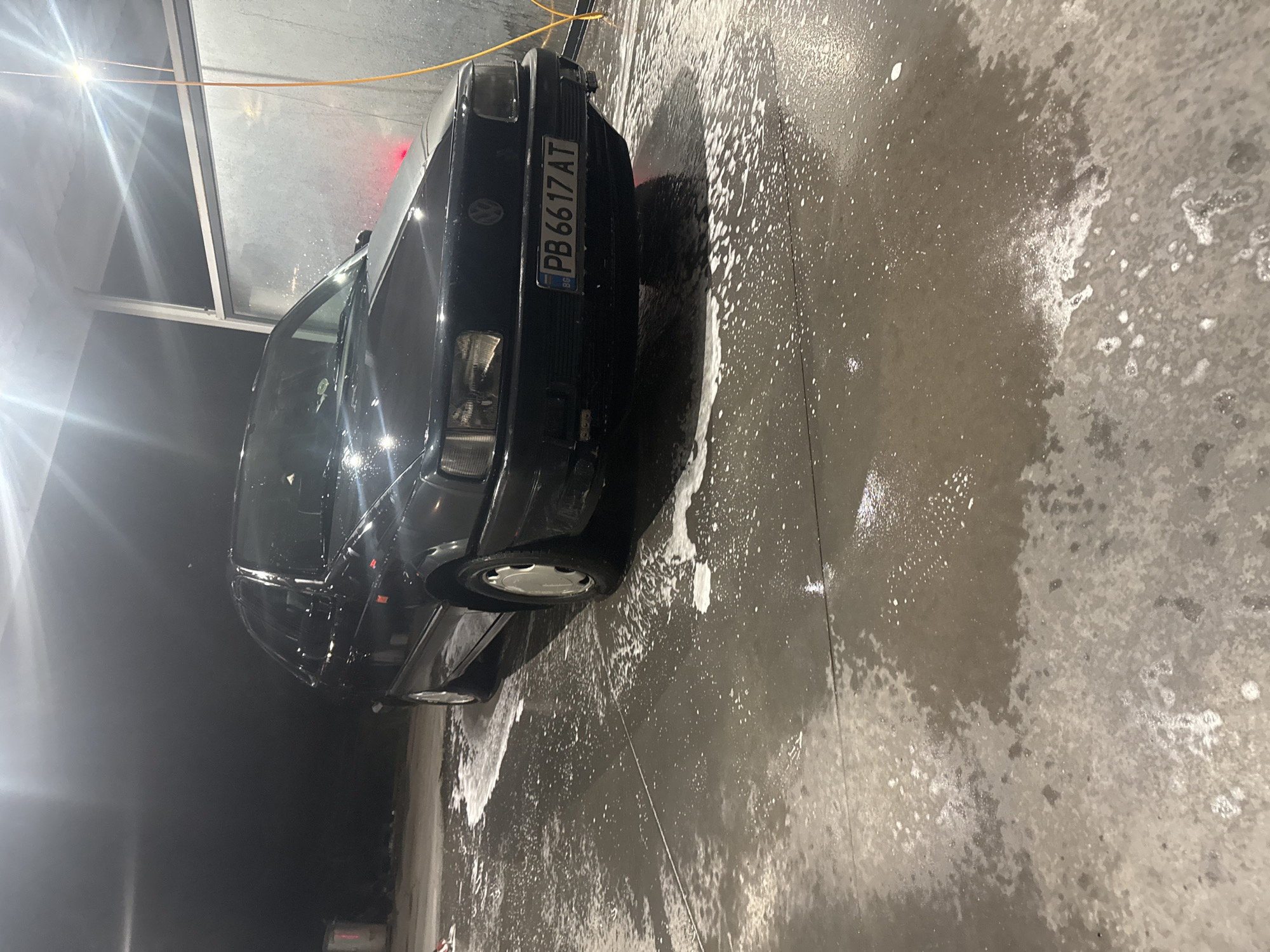 The image shows a black car, possibly a Volkswagen, being washed. The car's license plate reads "PB 6617 AT," indicating it is registered in Bulgaria. The setting appears to be an indoor car wash facility, given the presence of soap residue and concrete surfaces. The photo was likely taken in Plovdiv, Bulgaria, based on the license plate. There are no visible people in the image. The environment appears industrial.
