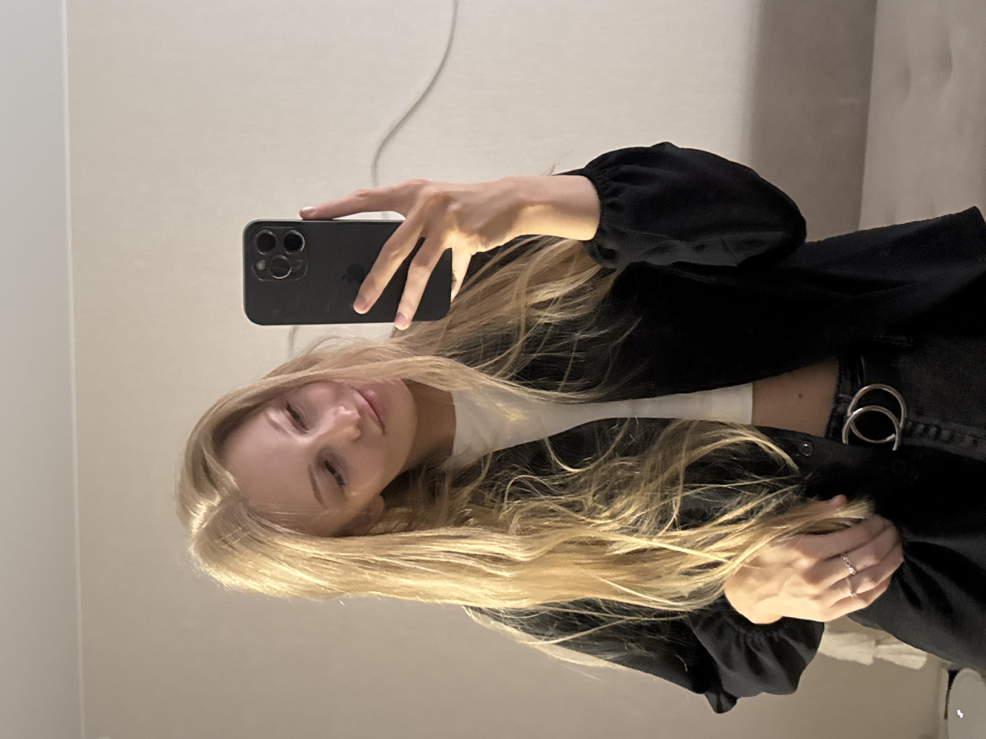 A young woman with blonde hair takes a mirror selfie indoors. She is wearing a black jacket over a white top and dark jeans. She is holding a black smartphone in her right hand, which is reflecting her face. She is wearing two rings on her left hand. The background appears to be a wall and a couch. The image was taken in the city of Perm, Russia.