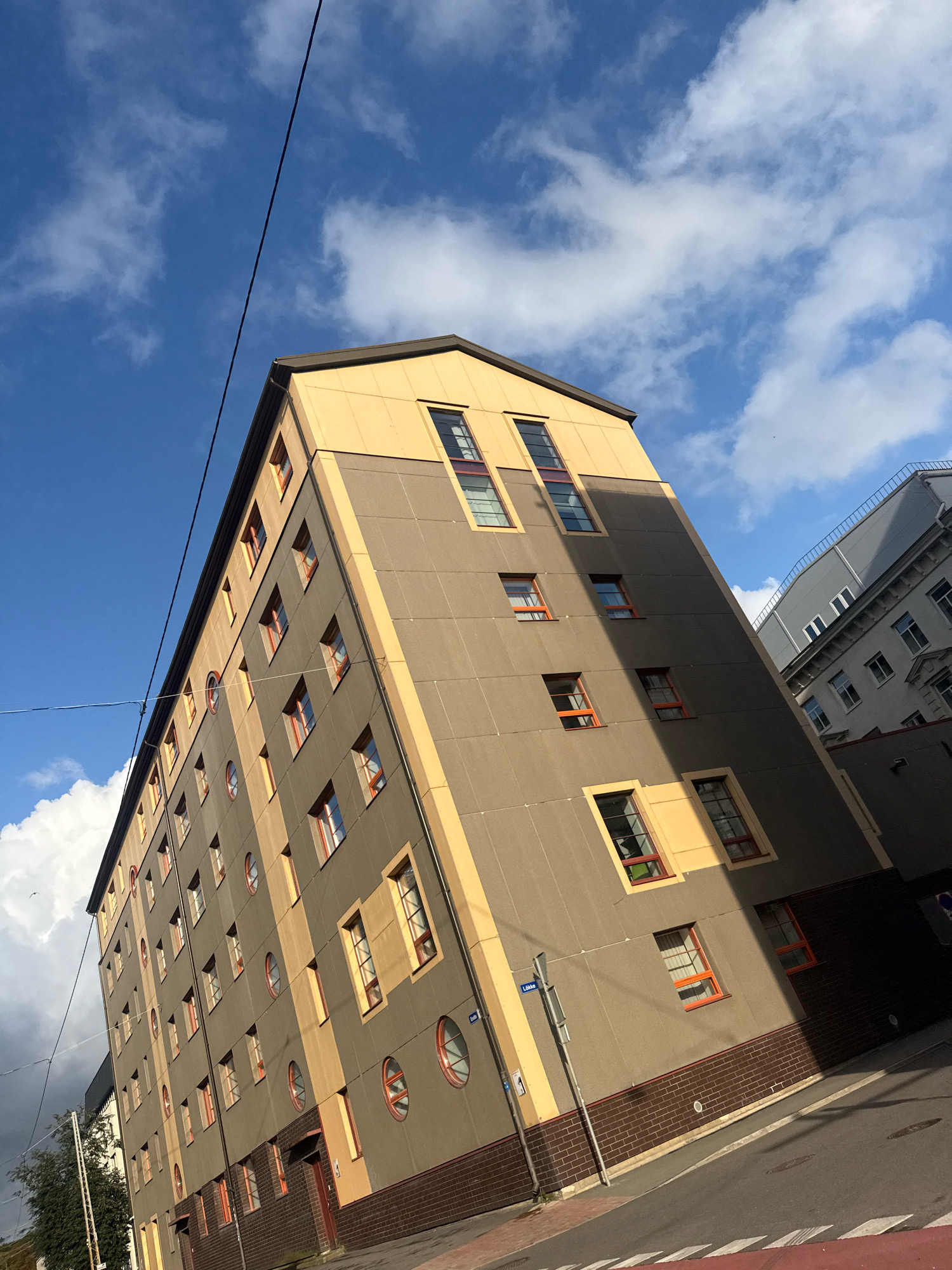 The image depicts the side of a multi-story building with a distinctive modern architectural design. The building's facade is primarily a neutral gray, accented with cream-colored vertical bands and a cream-colored pitched roof. Numerous windows are visible, many with vibrant orange frames, breaking the uniformity of the facade. Some windows are rectangular, while others are circular, adding a unique visual element. The building is situated on a street with a paved road and sidewalk. In the background, another building with a more traditional facade and numerous windows is partially visible. The sky is a clear blue with scattered white clouds, suggesting daylight. A power line is visible diagonally across the upper portion of the image. Based on the street sign visible on the building, "Lõõtsa", the location is Lõõtsa Street in the Ülemiste district of Tallinn, Estonia. There are no people or animals visible in the frame.