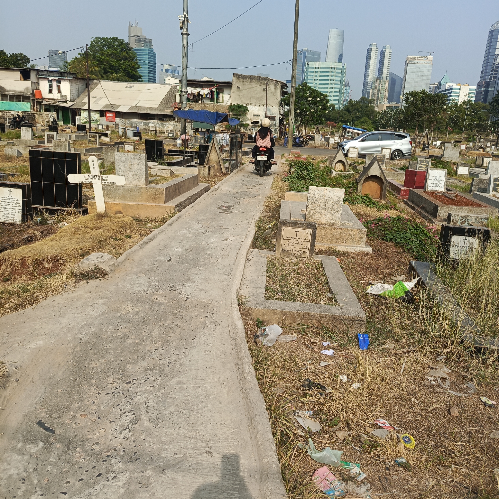 The image depicts a cemetery in South Jakarta, Indonesia. A paved pathway winds through the cemetery, bordered by numerous gravestones. Two individuals on a motorcycle, one driving and one as a passenger wearing a helmet, are moving along the pathway. In the background, several modern high-rise buildings are visible, contrasting with the cemetery's environment. The ground is covered with dry grass and scattered litter, including plastic bags and wrappers. Several gravestones have inscriptions, and some are adorned with flowers or crosses. The overall scene captures a juxtaposition of urban development and a place of rest.