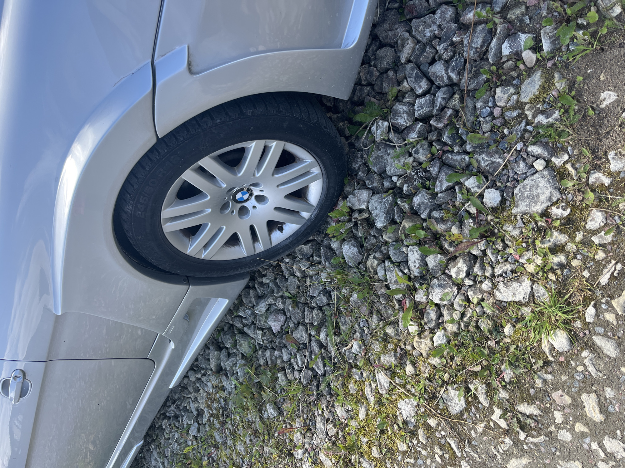 The image shows a close-up view of the rear passenger side of a silver BMW car parked on a gravelly surface with sparse green vegetation. The car's wheel, tire with visible "245/50 R 18" markings, and a portion of the fender and side skirt are visible. The car's bodywork appears to have some custom body kit elements, including flared fenders and aerodynamic side skirts. The ground is composed of various sizes of grey stones and dirt, with small clumps of green grass and weeds growing between the rocks. The lighting suggests it is daytime, with some reflections visible on the car's surface. There are no people or visible actions occurring in the scene. The location is described as being in the city of Il'ino, Russia.