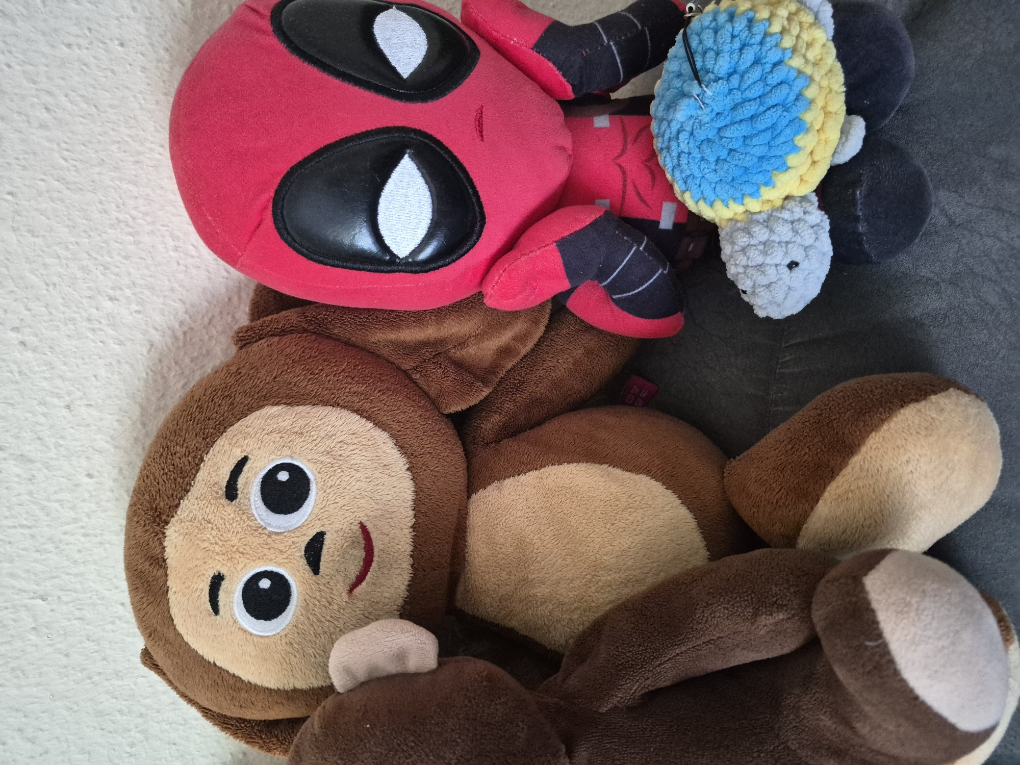 A top-down, close-up shot shows a collection of plush toys. In the foreground, a brown monkey plush toy with large, round black eyes and a smiling red mouth is lying on its side. The monkey has light brown fur on its face and belly, with darker brown fur on its arms and legs. Partially overlapping the monkey is a red and black plush toy resembling Deadpool. This toy has large white eyes with black rims, and red and black limbs. Attached to the Deadpool toy is a crocheted plush toy that appears to be a turtle or similar creature. This crocheted toy has a blue and yellow shell, and a light gray head with small black eyes. The toys are resting on a textured gray surface, with a white fuzzy surface visible on the left side of the frame. No specific time of day, weather, or prominent location cues are discernible from the image. There is a small pink tag with some text on it visible on the Deadpool toy, but the text is too small to read.