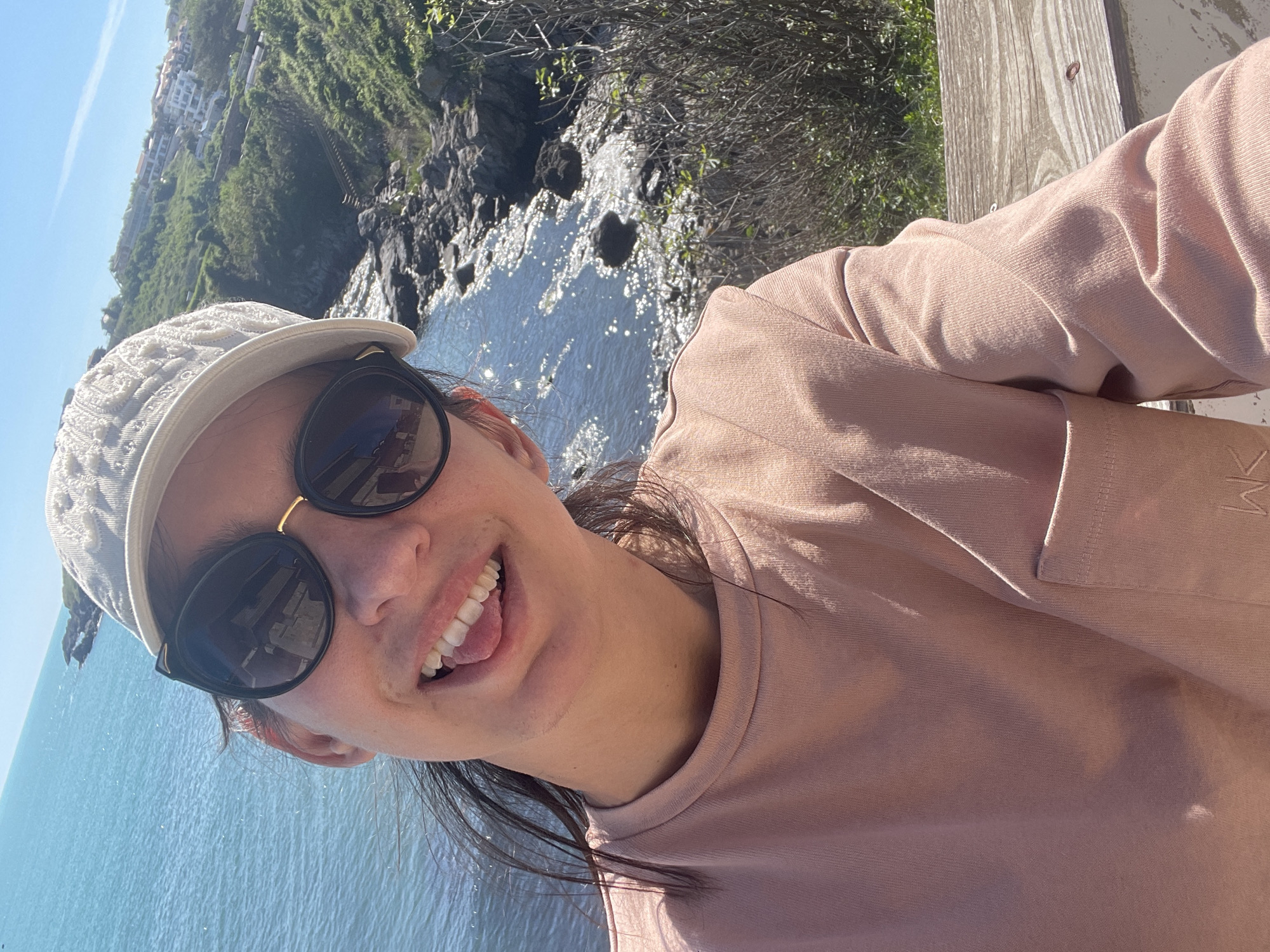 The photograph is a selfie of a person wearing sunglasses, a white cap, and a pink long-sleeved shirt. The individual is smiling with their tongue slightly protruding. The background features a view of the Black Sea, a rocky coastline, and a hillside with buildings, suggesting a coastal setting.
The location is likely in Sozopol, Bulgaria, based on the presence of coastal features, similar to the Black Sea coastline, and the hillside architecture. The person is likely on a wooden deck or platform.