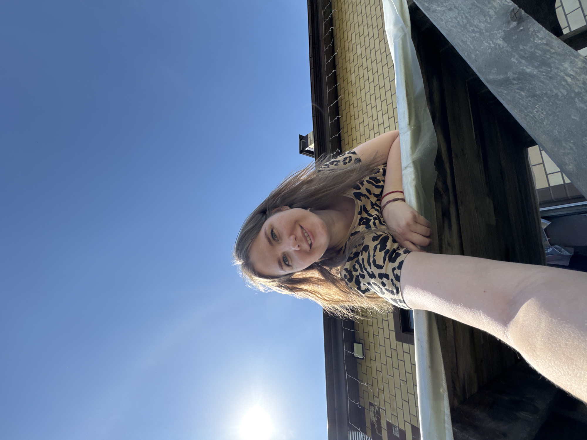 A low-angle, full-daylight photograph captures a woman smiling at the camera, leaning out from what appears to be a multi-story building or structure. The woman has long, light-brown hair and is wearing a brown and black leopard-print top, and at least one red bracelet on her left wrist. Her right arm is outstretched towards the camera. The background comprises a clear blue sky and a building facade constructed of light-yellow brick. A grey metal structure partially frames the scene, suggesting the vantage point might be from a balcony or elevated walkway. Given the location mentioned in the prompt, this scene appears to be in Ферма, Russia.