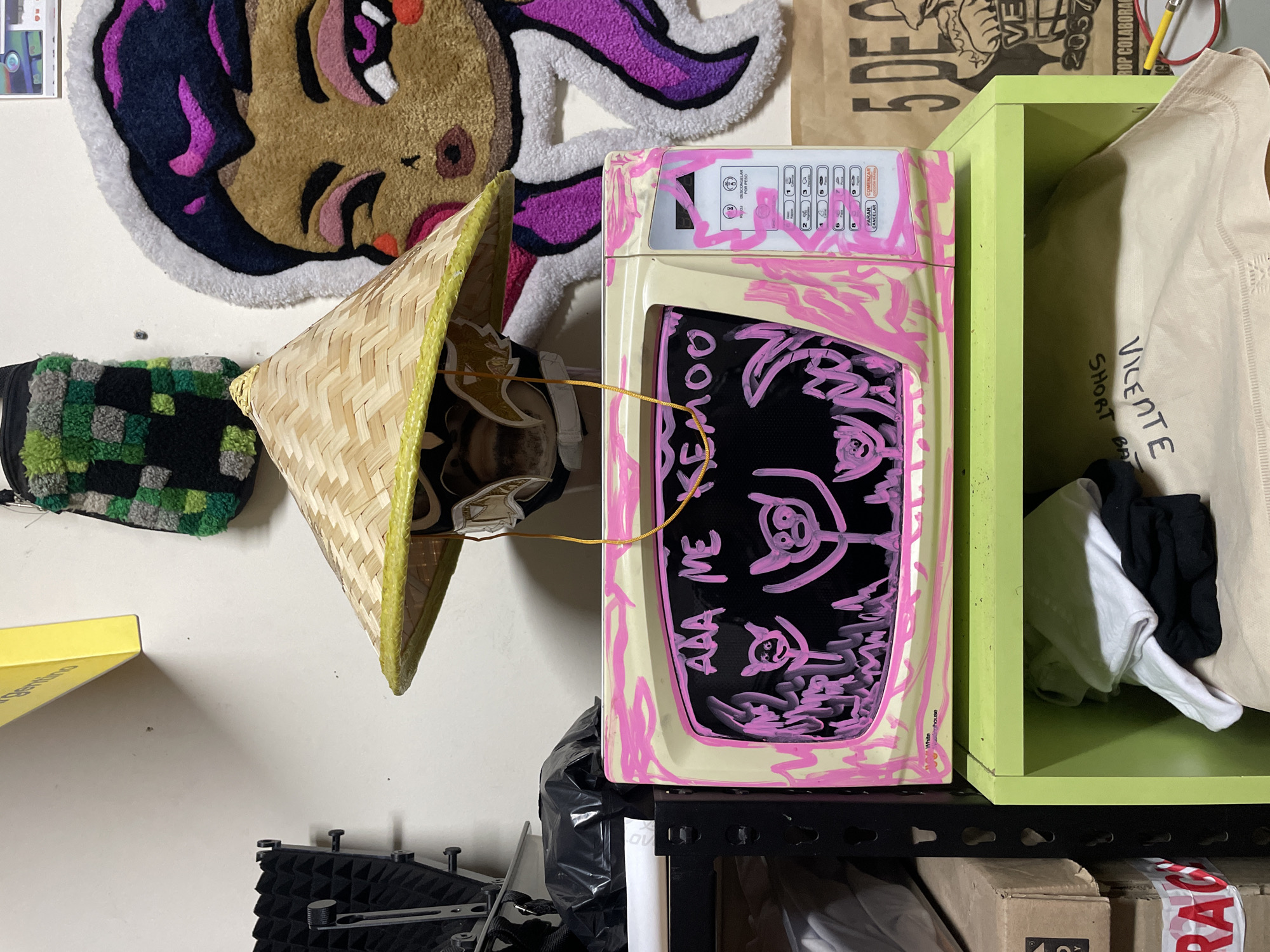 The image presents an assortment of objects and textures against a white wall. On the upper left, there's a round rug depicting a face, followed by a Vietnamese conical hat hanging next to a green and black checkered item. To the right, a small, beige appliance, possibly a microwave, is decorated with pink paint or marker, featuring abstract shapes and writing. The microwave sits atop a black plastic bag on a black wire shelf. To the right of the microwave, a bright green shelf unit contains a light-colored fabric tote bag, alongside some black and white fabrics or clothes. Beneath the shelf, there are cardboard boxes, one marked with "FRAGILE." In the lower left corner, part of a soundproofing panel is visible.
The appliance's control panel includes buttons in Spanish with the words "PARAR COMENZAR" and "POR PESO," suggesting the appliance is designed for the Latin American market, specifically mentioning Buenos Aires, Argentina. The text scrawled on the microwave appears to read "AAA ME KEMO0."