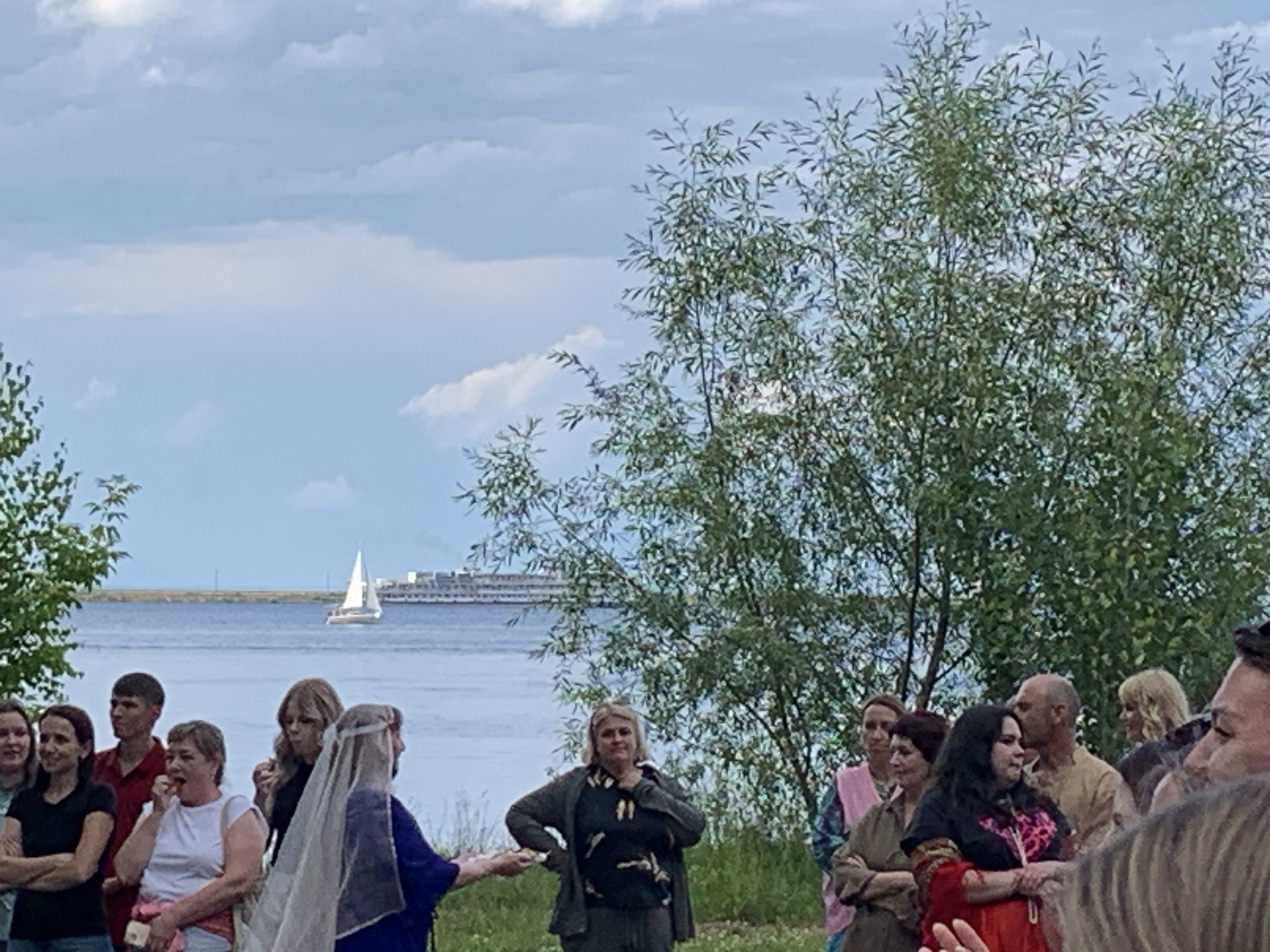 This media file depicts an outdoor gathering of people near a body of water, likely a river or lake, with a large vessel and a sailboat visible in the distance. The sky is overcast with scattered clouds, suggesting a moderate temperature and possibly a threat of rain. The scene is set in a grassy area with trees providing a natural backdrop.
Several individuals are the central focus of the image, appearing to be engaged in some form of communal activity or discussion. One woman in the foreground is wearing a white veil over her head and a blue robe, hinting at a possible cultural or historical reenactment or celebration. Another woman in a dark patterned top is looking towards the camera with a neutral expression. To her left, a woman in a white top is holding something to her mouth, possibly food. A man in a red shirt is standing behind her, looking to the side. Other individuals are visible in the background, some appearing to converse.
The overall atmosphere seems relaxed and informal. The time of day is difficult to ascertain precisely due to the overcast conditions, but it appears to be daylight hours, possibly late afternoon or early evening. There is no visible text in the image. The location context of Chaikovsky, Russia, suggests this event may be related to local customs or a community gathering by the Kama River.