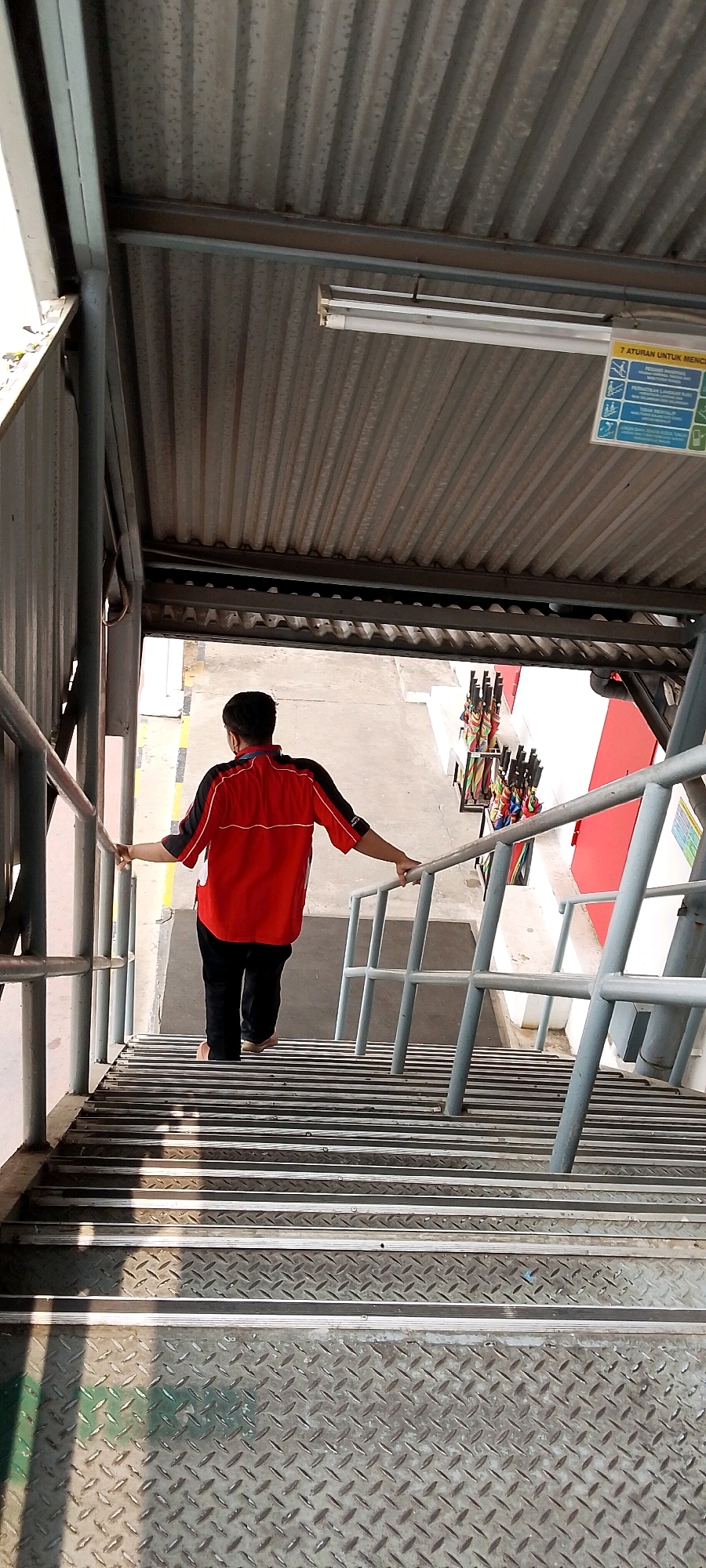 A person wearing a red and black shirt and black pants walks down a metal staircase under a corrugated metal roof. The staircase has metal railings on both sides and the steps are made of ridged metal. There are several colorful umbrellas in a stand to the right of the stairs, and a sign with text in blue and white is attached to the ceiling. The scene appears to be an outdoor or semi-outdoor area, possibly a public building or transit station, in Depok, Indonesia.