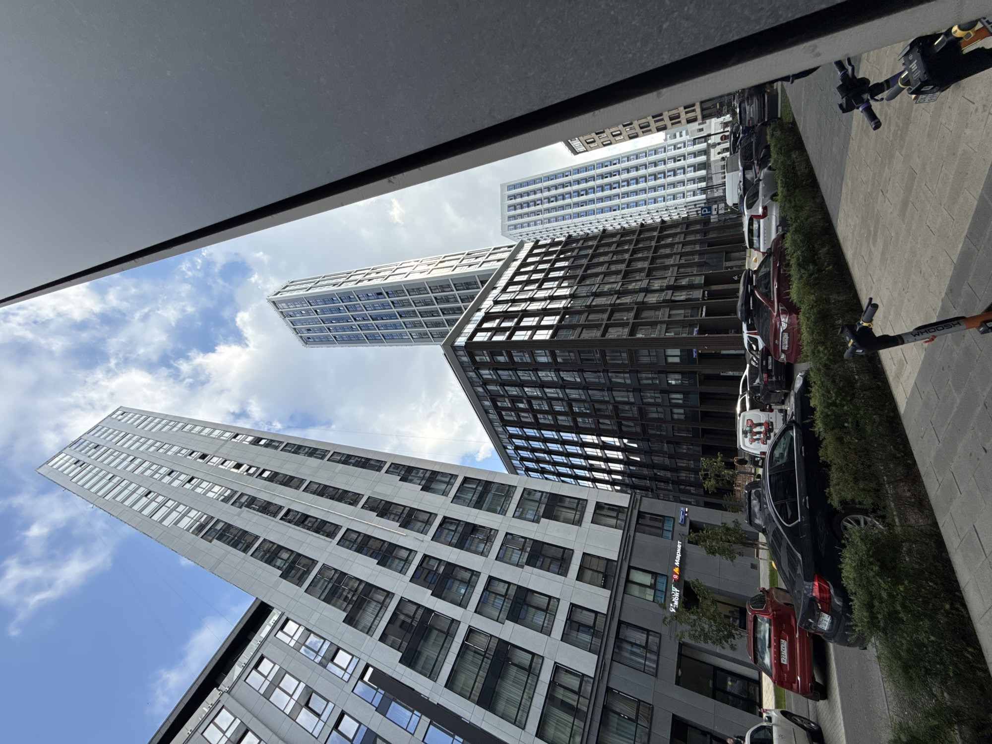 The image depicts a street scene in Moscow, Russia, characterized by modern high-rise buildings and parked cars. The perspective is upward-looking, emphasizing the scale of the architecture. In the foreground, a dark gray building facade partially obscures the view.
The central focus is on a collection of contemporary residential towers with distinct architectural styles. The leftmost building is a tall, slender structure with a light gray facade featuring numerous rectangular windows arranged in a grid pattern. To its right, a dark brown, cube-like building stands, also with a repeating window pattern. Further in the background, two more high-rise buildings are visible: one is a very tall, thin tower with a blue-tinted glass facade, and another is a white building with a similar grid of windows.
Below the buildings, on the street level, several cars are parked. A prominent red sedan is parked on the right side of the street, facing away from the viewer. To its left, a black sedan is parked. Other vehicles, including white vans and SUVs, are also visible. Two electric scooters are parked on the sidewalk in the lower right portion of the frame.
The sky above is a mix of blue with scattered white clouds, suggesting a daytime scene. Trees and bushes line the street and sidewalk, adding a touch of greenery to the urban environment. Signs are visible on the ground floor of one of the buildings, including one with Cyrillic text that appears to be a store name. The overall impression is one of a modern urban district with new construction.