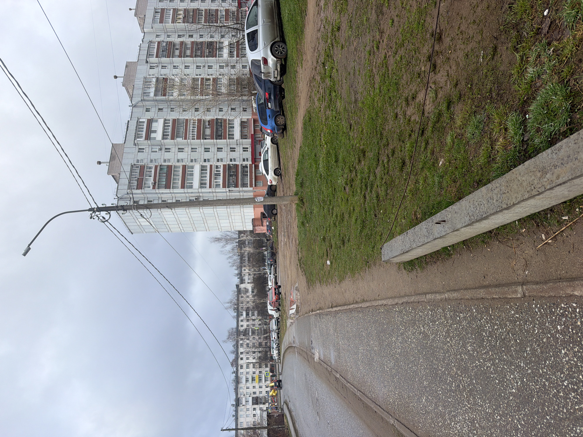 This image depicts an urban residential street in Chaikovsky, Russia, under an overcast sky. A large, multi-story apartment building, featuring numerous windows, balconies, and visible air conditioning units, dominates the left side of the frame. Its grey facade suggests typical Soviet-era construction.
In the foreground and extending into the mid-ground, a paved road or sidewalk borders a wide verge of sparse green grass and exposed muddy patches, indicative of a damp environment. Several cars are parked on this grassy area, including white, blue, and silver vehicles. Additional buildings and parked cars can be seen in the distance, lining the street.
A utility pole, equipped with a street light and multiple overhead wires, stands in the mid-ground on the left, with its wires stretching across the muted, cloudy sky that fills the upper portion of the image. In the immediate right foreground, a rectangular concrete slab lies horizontally on the grass, bearing faint, unreadable Cyrillic text or markings on its surface.
The scene appears to be set during daytime, but the heavy cloud cover gives it a dull, subdued atmosphere. No people are visible, and no particular activity or event is occurring, portraying a quiet, everyday snapshot of an urban neighborhood.