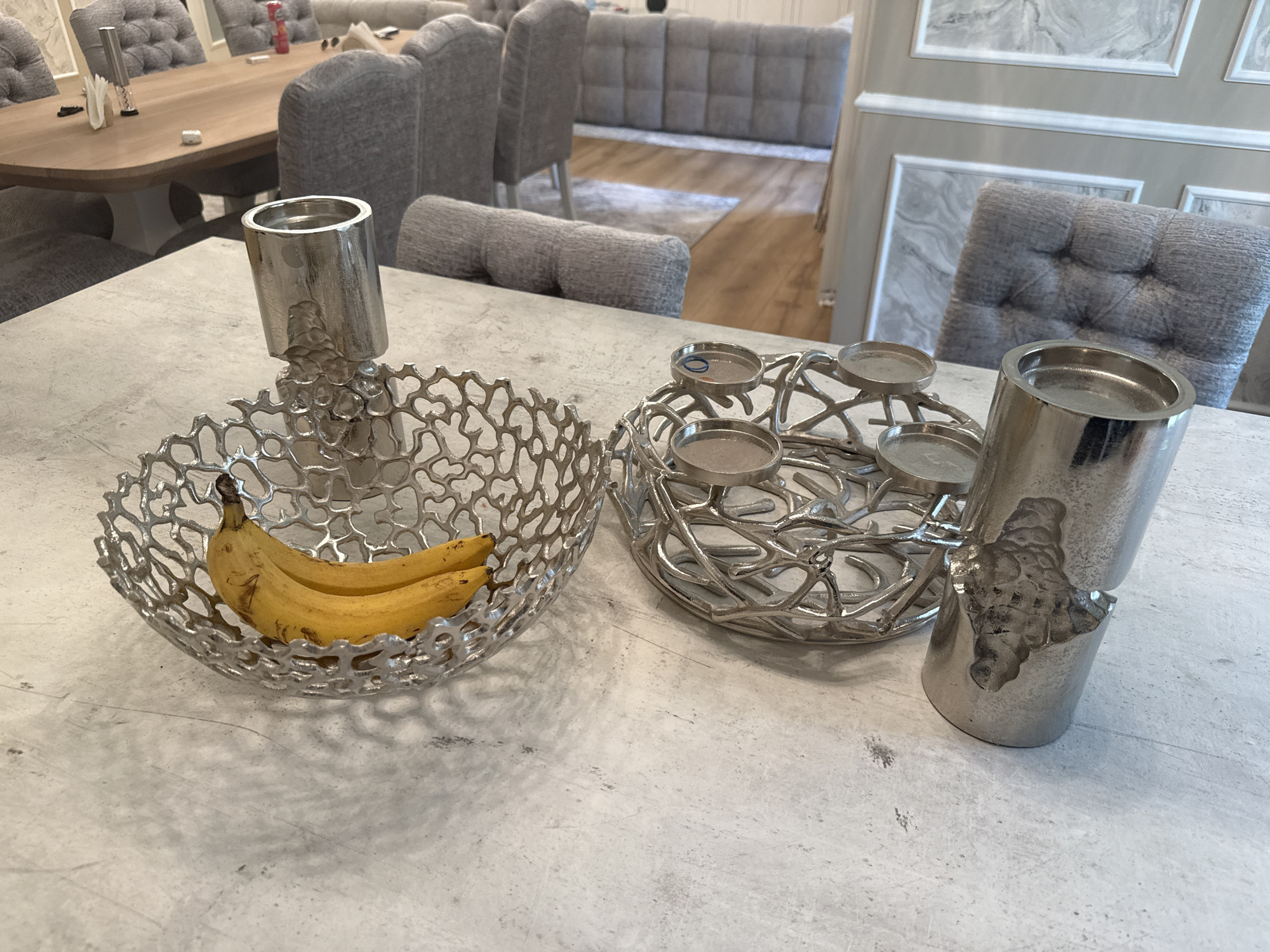 The image shows a close-up of a table setting, likely indoors. Two ornate silver-colored decorative objects are prominently placed on the table. One is a bowl-shaped metal basket holding two bananas. Adjacent to it is a multi-tiered silver metal candle holder. The table has a light grey surface with subtle patterns. In the background, out of focus, are upholstered chairs and a sofa, suggesting a dining or living room area. The style of the furniture is modern and upscale. There is no specific information to determine the location of the photo in Sofia, Bulgaria.