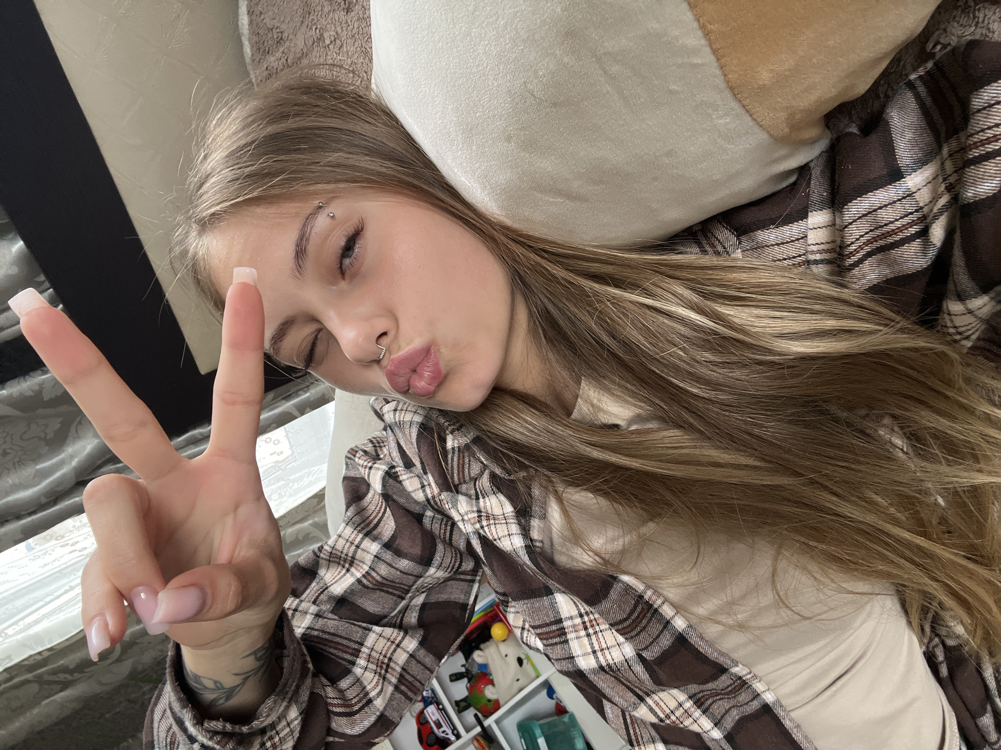 A young Caucasian female with long, blonde hair is lying on a light-colored surface, possibly a sofa or bed, and looking at the camera. She is wearing a brown and white plaid shirt over a light-colored top. Her right hand is raised, with her index and middle fingers extended in a peace sign, while her left hand is not visible. She is winking her left eye and puckering her lips. She has a nose ring and an eyebrow piercing. Behind her, there is a white pillow and a curtain with a textured pattern. To the left of the frame, a shelf unit with several compartments is visible, containing toys and other objects. The scene is indoors and appears to be a residential setting. The specific location in the city of Фоки, Russia cannot be determined from the visual information provided in the image.