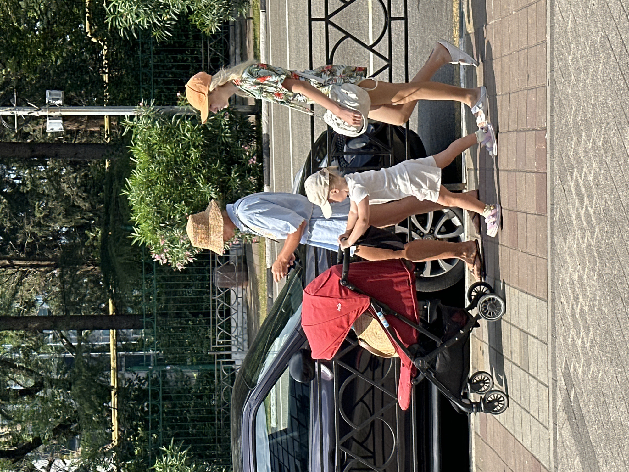 The image depicts three people and a stroller on a sidewalk next to a street and a black car. A woman with blonde hair and an orange hat is walking on the sidewalk, wearing a floral print shirt and white shorts. She is holding a white bag. Beside her, a man wearing a blue shirt and a straw hat is leaning over, appearing to assist a small child. The child is wearing a white outfit and a light-colored cap, and is holding onto the stroller. The stroller is red and black and has a straw hat inside it. The sun is casting shadows, suggesting it is a sunny day. The background includes greenery and a decorative black metal fence. The overall scene suggests a family outing on a pleasant day, possibly in a residential or park area. Visible text on the stroller reads "JOIE". The image was taken in Sochi, Russia.