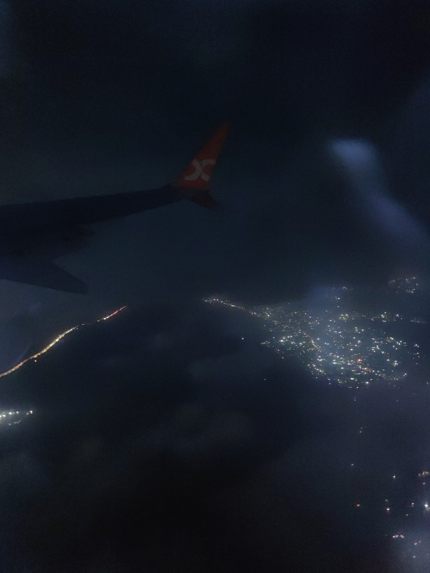 The image is an aerial view of a city at night, likely taken from an airplane. The foreground features a portion of an airplane wing, identifiable by its dark color and a red logo with a stylized 'X' on its tail. Below the wing, city lights are visible, scattered across the dark landscape, suggesting a densely populated area. The lights form clusters and streaks, indicating roads and buildings. Some lights appear blurred due to the movement of the aircraft or atmospheric conditions like clouds or fog. The scene is dominated by darkness, with the city lights providing the only significant illumination. The location is identified as Panvel, India.