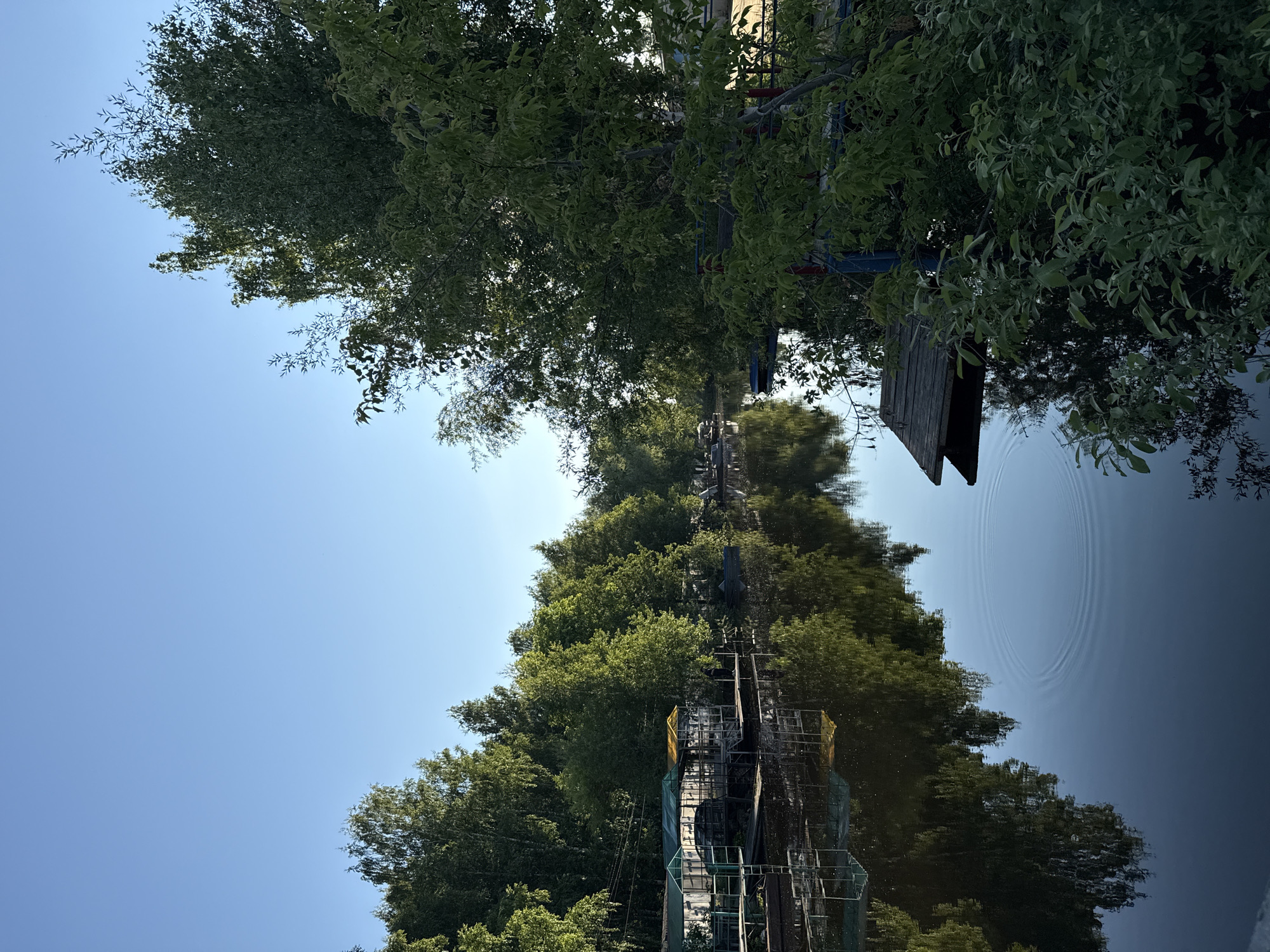 A serene outdoor scene is captured, featuring a body of water bordered by a lush treeline under a clear blue sky.
**Subjects:**
The primary subjects are dense green trees, a calm body of water, and a wide expanse of clear blue sky. Along the treeline on the left bank, several man-made structures are partially visible. These appear to be metal frameworks, possibly scaffolding or parts of a water-related installation, with one section covered in yellow material. On the far right bank, a wooden structure, possibly a small dock or shelter, extends towards the water.
**Activity or event taking place:**
No specific activity or event is explicitly depicted. The scene is static, showcasing nature. However, concentric ripples are visible on the water's surface, suggesting a recent, unseen disturbance.
**Notable details:**
The time of day appears to be during daylight hours, likely mid-morning to early afternoon, given the bright, clear blue sky and the even illumination. The weather is sunny and clear. The calm water acts as a mirror, providing clear reflections of the trees and sky, contributing to the tranquil atmosphere. The combination of natural elements with the subtle presence of man-made structures suggests a location where nature and human intervention coexist, possibly a developed natural area like a park, riverbank, or lake.
**Visible text:**
There is no visible text in the image.