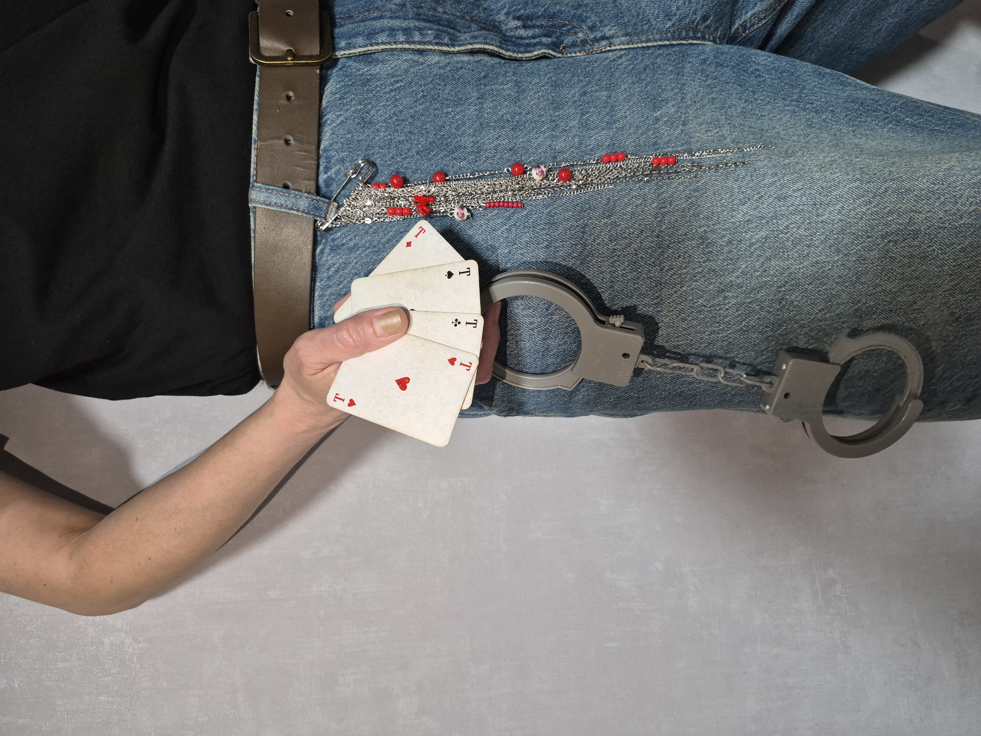 A medium shot shows a person holding four playing cards in their right hand and a pair of handcuffs hanging from their belt loop. The person is wearing a black t-shirt, a brown leather belt, and blue denim jeans. The playing cards appear to be Aces, all with red hearts. A silver chain with red and pink beads hangs from the belt loop on the right side of the image. The background is a plain, light gray textured wall. The scene is indoors.