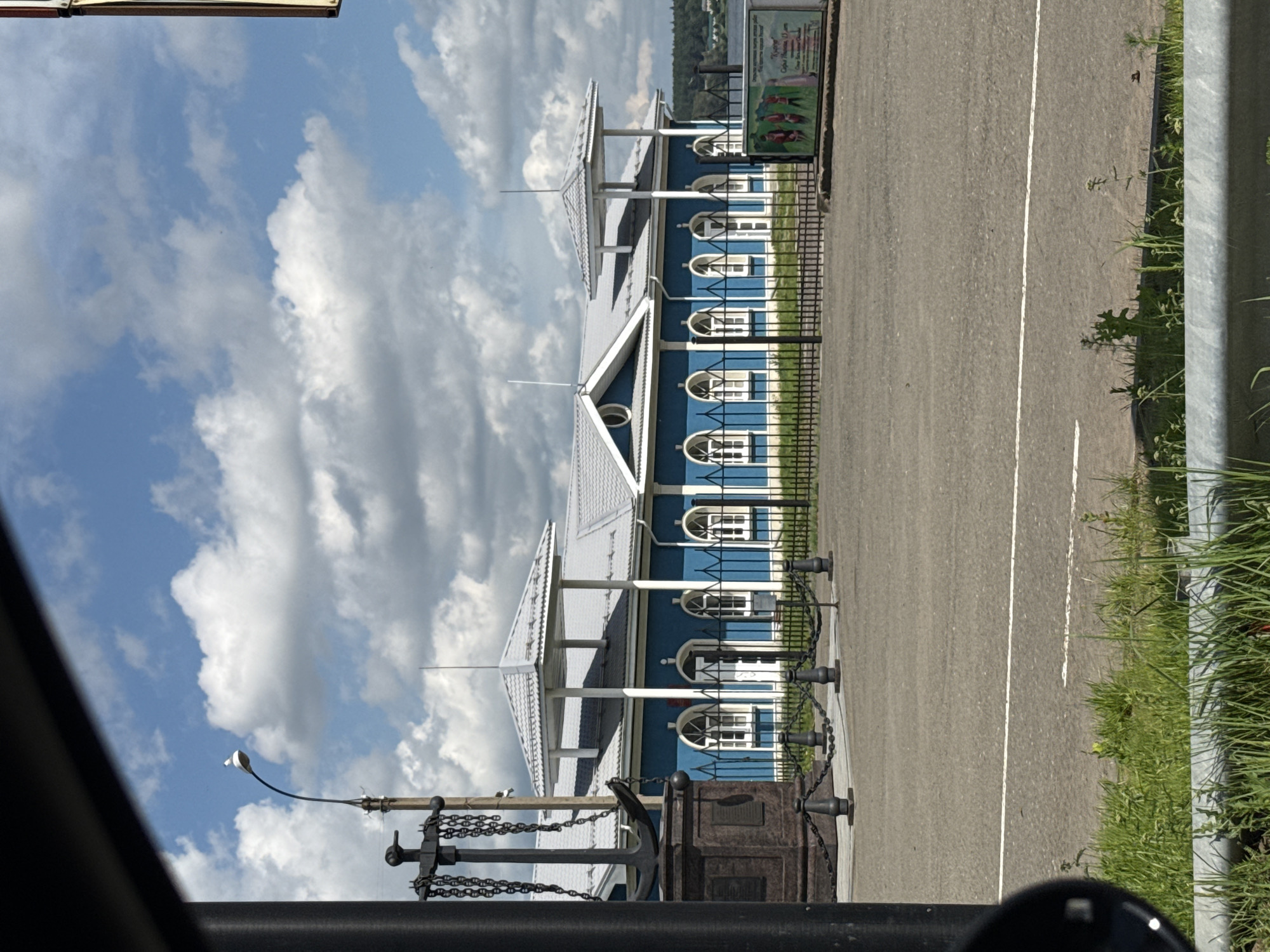 The image shows a blue building with a white roof and a covered walkway, likely part of a historical or cultural attraction in Votkinsk, Russia. The building has many arched windows with white frames. In front of the building, there is a black metal fence with bollards and chains. An anchor is prominently displayed near the fence. The scene is set on a sunny day with some fluffy white clouds in a bright blue sky. A paved road with white lines runs alongside the building and fence, with a strip of green grass and weeds bordering it. On the far left, a black object, possibly a car mirror, is visible, suggesting the photo was taken from a vehicle. In the background, a sign with text and images is attached to the fence, and a distant tree-covered hillside can be seen. The overall mood is calm and bright, suggesting a pleasant day for visiting the location. The time of day appears to be midday, given the bright sunlight.