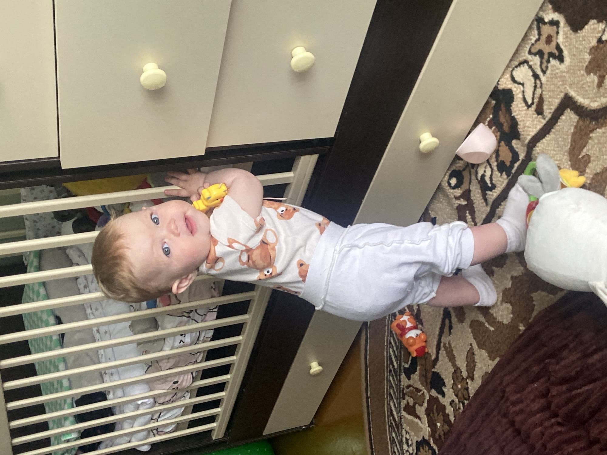 A baby lies on their side, partially in an open drawer of a dresser, with a yellow toy in their hand. The dresser is two-toned, with a light top portion and a dark wood-colored bottom section. A crib with patterned bedding is adjacent to the dresser. On the floor, a plush toy and a small toy car sit on a patterned rug. The image was taken indoors in Фоки, Russia.