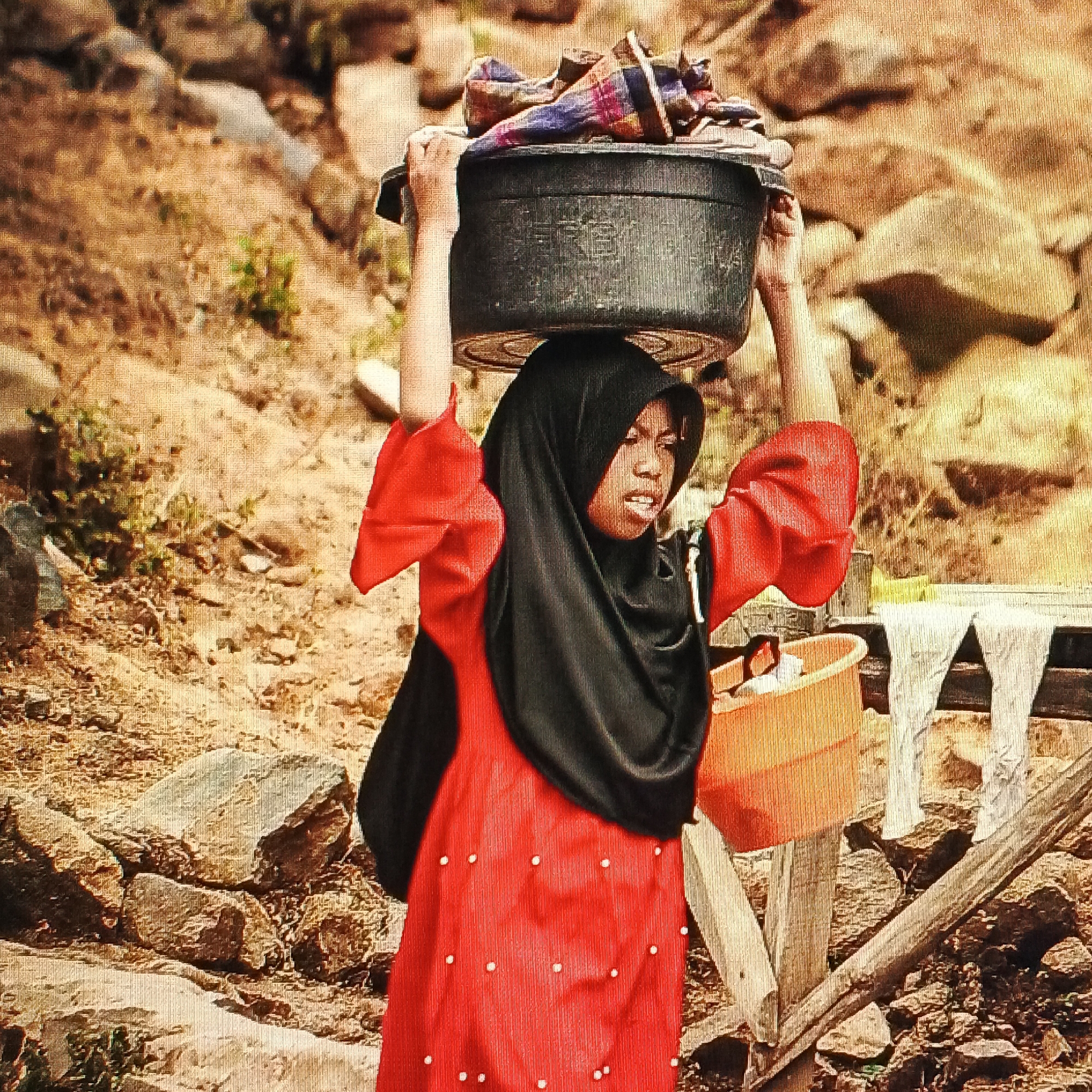 A young woman, wearing a red dress and a black hijab, carries a large black basin filled with clothes on her head. She has her hands raised to balance the basin. Beside her, to her right, is an orange bucket. The background is rocky and dry, suggesting an outdoor, possibly rural, environment. The image appears to be a photograph with a textured, slightly grainy filter applied.