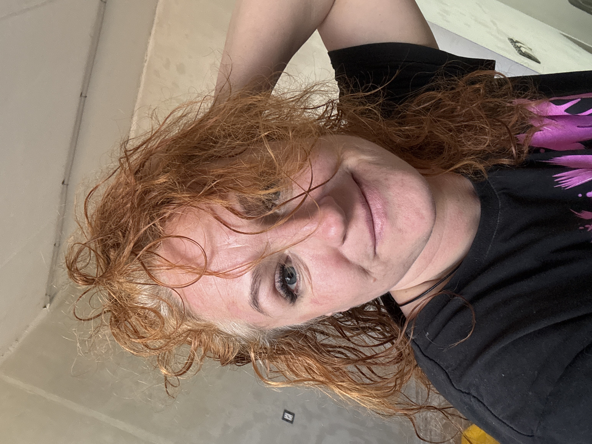 A portrait of a woman with red, curly hair. She is wearing a black t-shirt with a pink floral design. The background consists of a light-colored wall and a portion of a floor. The location of the city of Chaikovsky, Russia is unknown.