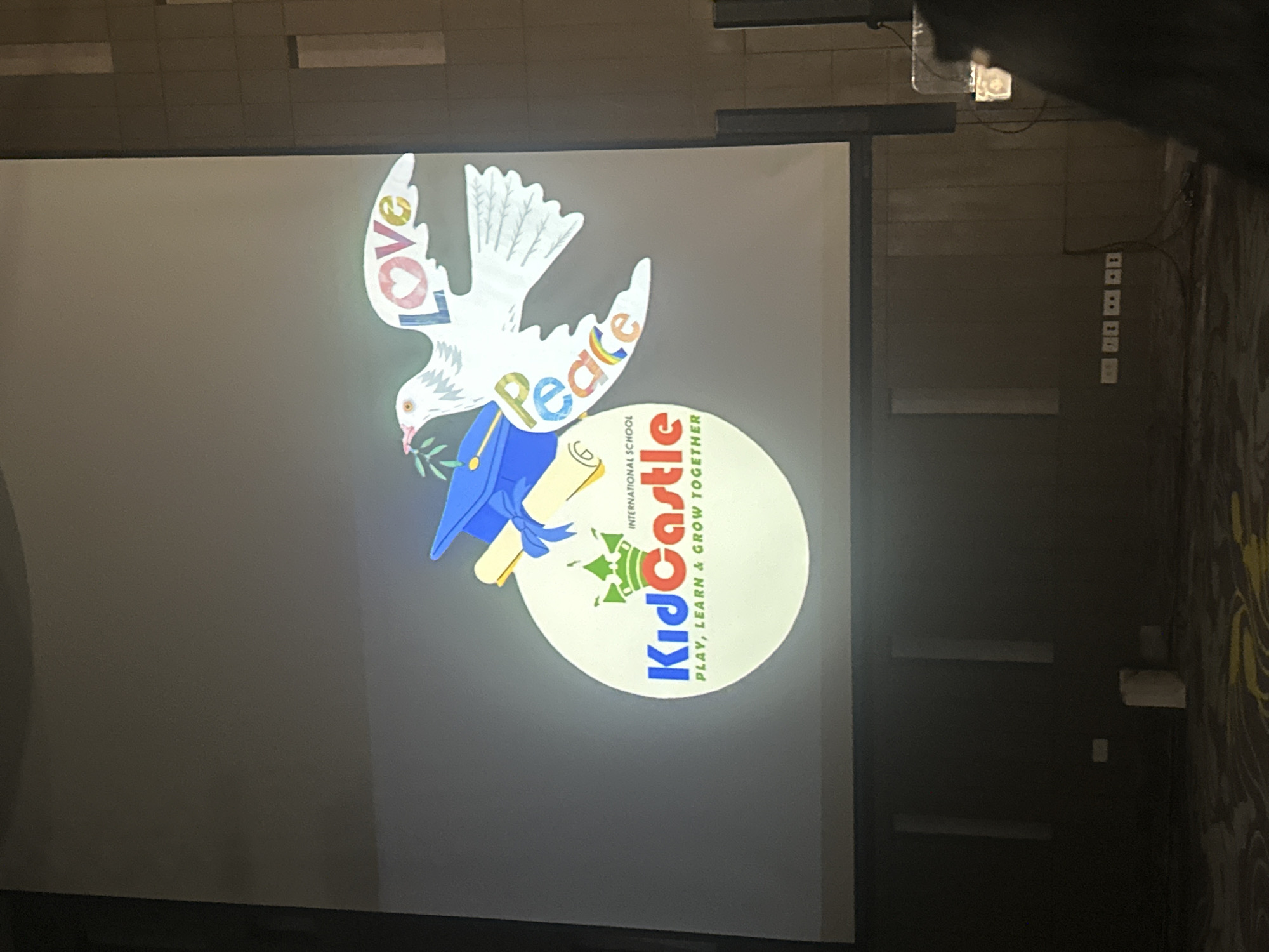 The media file displays a projection on a large screen, likely in an indoor setting such as a school auditorium or conference room in Nha Trang, Vietnam. The projection features a graphic with a dove holding an olive branch in its beak, symbolizing peace. The dove is white with intricate feather details and its wings are spread wide. The words "Love" and "Peace" are creatively integrated into the dove's wings, with "Love" written vertically in colorful lettering and "Peace" also in colorful lettering arching over the dove's body.
Below the dove, a circular logo is visible, which appears to be for "KidCastle International School". Inside the circle, the text "KidCastle" is prominently displayed in large blue and red letters. Beneath "KidCastle", the tagline "PLAY, LEARN & GROW TOGETHER" is written in smaller text. The logo also includes a depiction of stylized green trees. The overall tone of the graphic is cheerful and educational, conveying a message of positivity and growth.
The surrounding environment visible in the background suggests a dimly lit room, with the screen being the primary light source. There are some electrical outlets and wall fixtures visible on the right side of the frame, indicating a functional interior space. The image is taken from an angle that suggests the viewer is looking up at the screen. The time of day is not discernible from the image, and no weather conditions are apparent. The overall impression is of a school event or presentation.