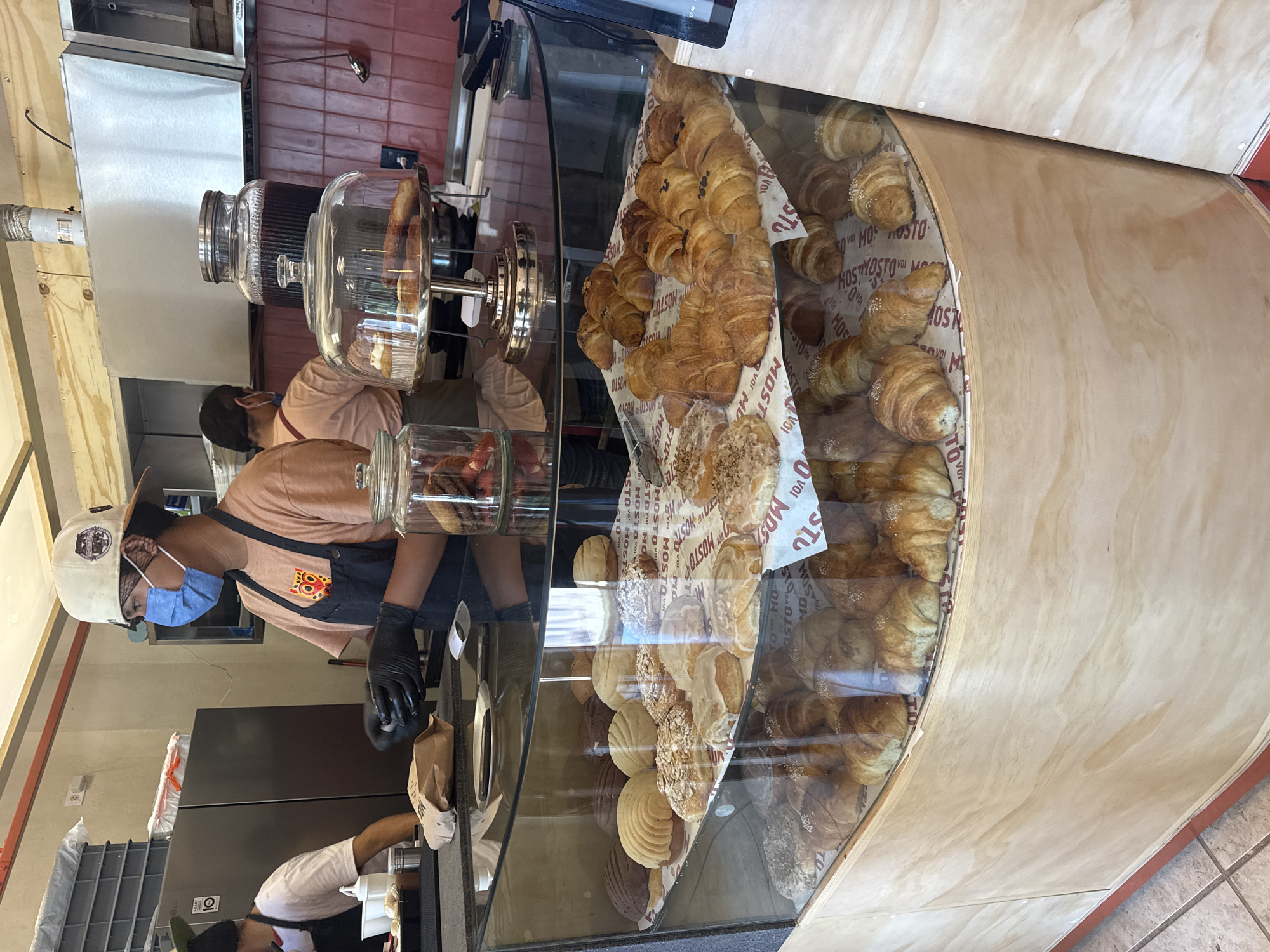 Here is a description of the image.
The image shows a bakery setting, possibly in San Andrés Cholula, Mexico. A display case is prominently featured, filled with an assortment of baked goods, including croissants and conchas (Mexican sweet bread). A person wearing a blue face mask, a peach-colored shirt with a logo, black gloves, and a hat is behind the display case, interacting with the items. The person appears to be using tongs or similar utensils to pick up an item, possibly placing it into a brown paper bag. The display case is constructed with glass and a light-colored wood. Background elements include kitchen equipment, shelves, and parts of the bakery interior. The writing on the bakery paper insert is "MOSTVO" and is likely a bakery brand name.