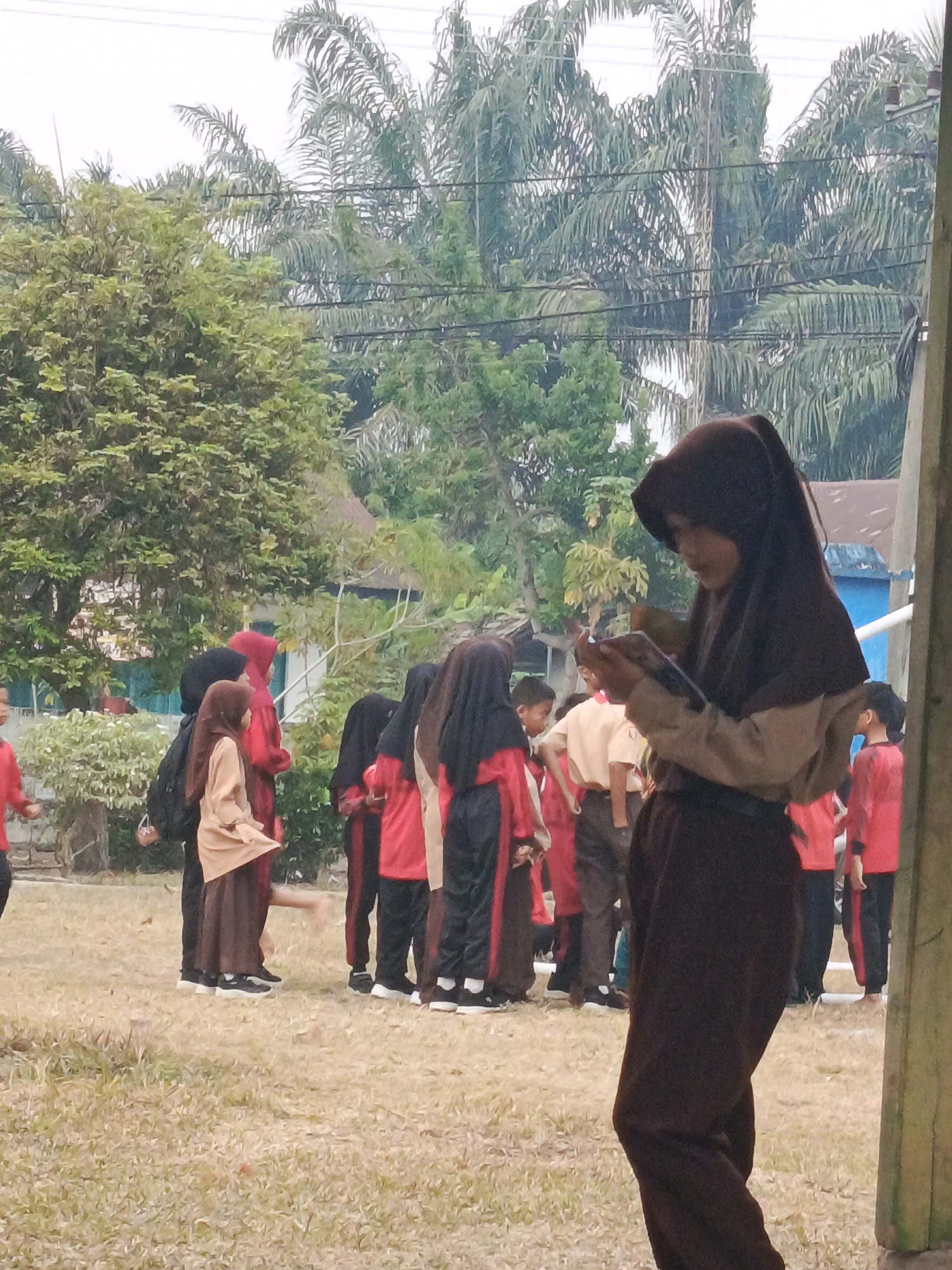 The image shows a group of young people, likely students, outdoors in a grassy area with trees and buildings in the background. The person in the foreground, wearing a dark brown hijab and uniform, is looking down at a handheld device. Several other individuals are visible in the middle ground, some wearing red and black uniforms with hijabs, and others in lighter-colored uniforms. The environment appears to be a schoolyard or public space, with a cloudy sky overhead. The location is identified as Megang Sakti II, Indonesia, suggesting a tropical or subtropical climate due to the presence of palm trees.