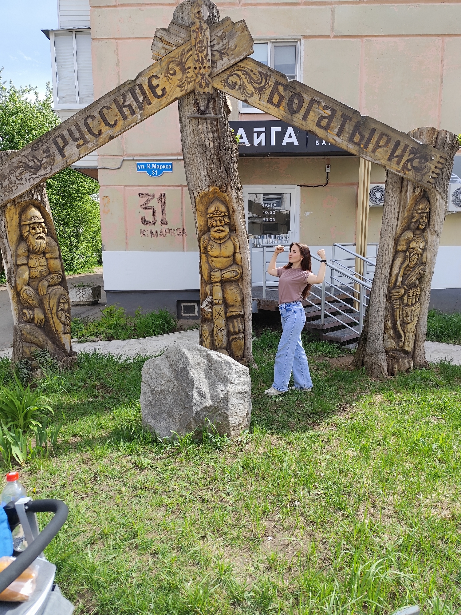 A sunny, outdoor scene depicts a young woman flexing her biceps in front of a wooden structure. The structure consists of three carved wooden pillars connected by two horizontal beams, forming an archway. The carvings on the pillars depict figures resembling ancient warriors or folk heroes. The beams bear the inscription "Русские Богатыри," translating to "Russian Bogatyrs" or "Russian Knights." In the background is a building with a sign that reads "ТАЙГА" (TAIGA), which is a Russian word referring to a type of biome. A small sign on the building provides the opening hours, from 10:30 to 20:30. The building is addressed as "ул. К. Маркса 31" (K. Marks Street 31). In front of the wooden structure is a large, grey boulder. The woman is wearing a brown t-shirt and light blue jeans. A stroller is partially visible in the lower left corner of the image. The location is identified as the city of Chaikovsky, Russia based on the Cyrillic inscriptions and common Russian street name.