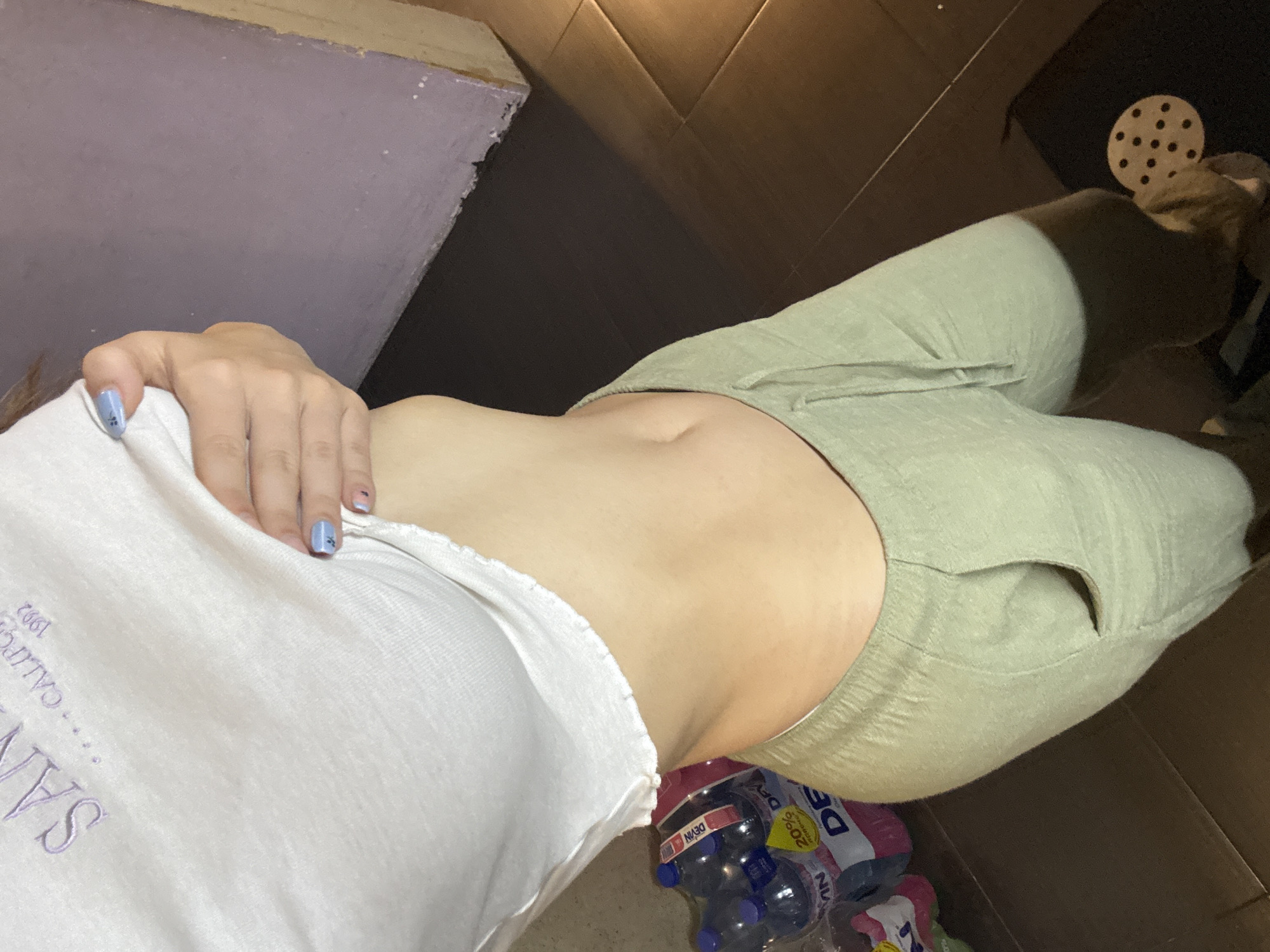 The image shows a person's midsection, focusing on their stomach and waist. They are wearing a white crop top and light green drawstring pants. The background includes a purple wall, dark wooden flooring, and a stack of plastic water bottles on the left. The overall lighting is dim. No specific location in Sofia, Bulgaria can be identified from the image.