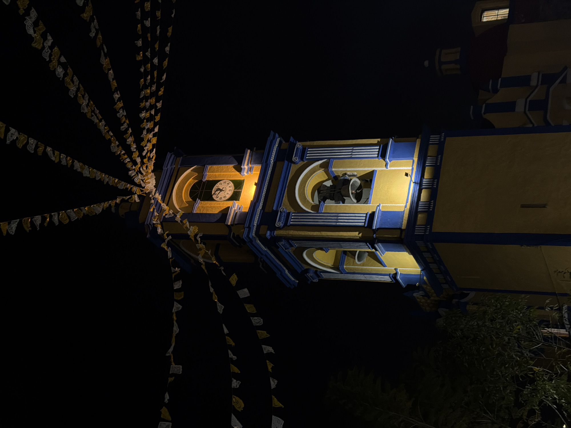 The image shows the illuminated bell tower of the Templo de San Gabriel in San Andrés Cholula, Mexico, at night. The tower is painted in yellow and blue, with a clock visible on one side. A bell is visible in one of the openings. Strings of decorative flags are strung across the tower, illuminated by the same light source. The background is dark, but other parts of the church building are visible at the top right of the frame. Some foliage is also visible at the bottom right.