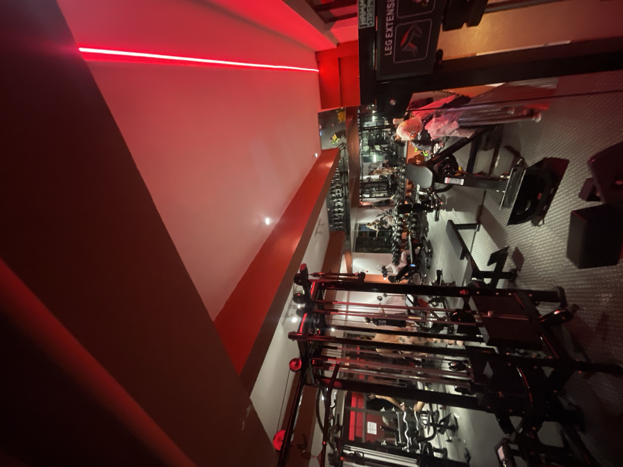 A gym interior is depicted with red lighting accents along the ceiling. In the foreground, a black multi-gym machine with cables is visible. To the right, a piece of fitness equipment labeled "LEG EXTENSI" is partially visible. The middle ground shows various weight machines and racks of dumbbells. Several individuals are engaged in workouts or resting on benches throughout the gym. The floor appears to be a dark, textured mat. The scene is lit with a combination of overhead and accent lighting, creating a dramatic ambiance. This image was captured in the city of Chongqing High-tech Zone, China.