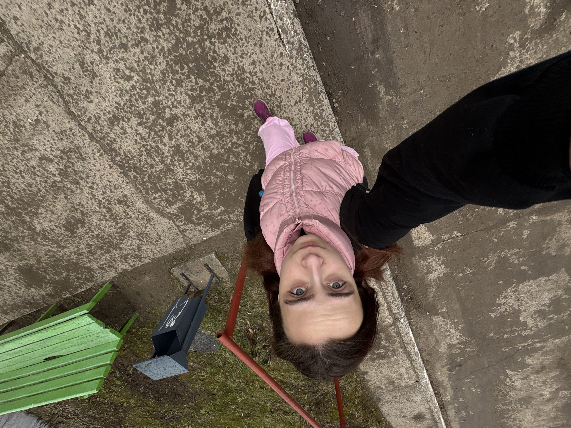 A young woman is captured in an outdoor, high-angle selfie in Chaikovsky, Russia. She is positioned towards the center-top of the frame, appearing upside down due to the camera's perspective, looking directly at the viewer with a neutral expression and light-colored eyes. Her brown hair is visible around her face. She is wearing a light pink puffer vest over a dark long-sleeved top and pink trousers, paired with magenta-purple sneakers.
The setting appears to be a public outdoor space, possibly a park or playground, with elements of urban infrastructure. The ground beneath her is a textured, mottled concrete or pavement, suggesting dampness or wear. To her left, partially visible, is a green wooden slatted bench. Further to the left and slightly below her is a dark gray or black metal bin, possibly a pet waste dispenser, featuring a white graphic or silhouette (resembling a person or animal with a bag) and some indistinct white text. Red metal bars, possibly from a fence or playground equipment, are in the foreground, framing parts of the scene. The ground near the bench and bin transitions to patchy grass and dirt.
The overall lighting suggests daytime, possibly overcast, as there are no harsh shadows. The woman's attire indicates cooler weather. The image focuses on the woman while providing context of her immediate surroundings.