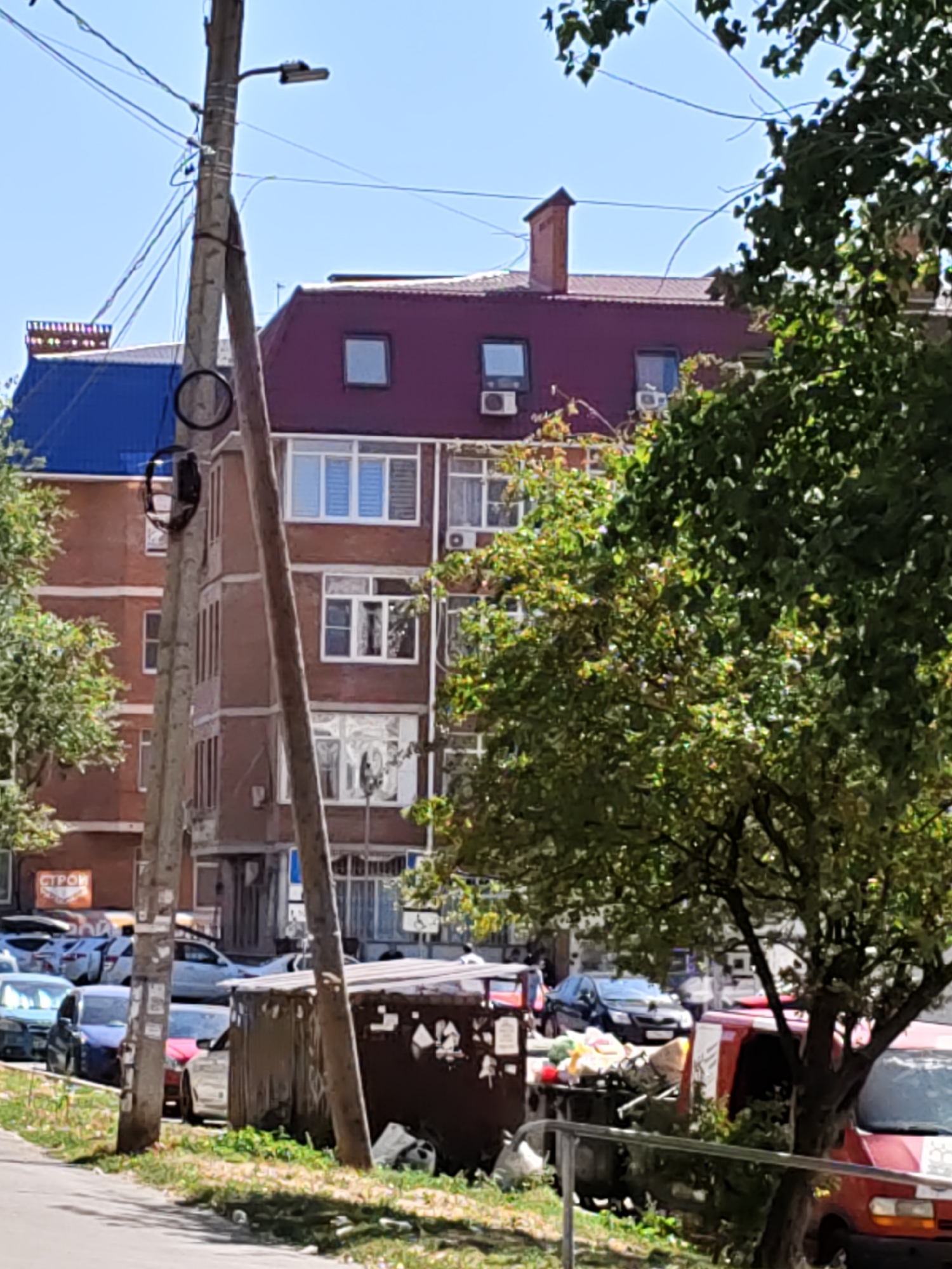 The image shows a street scene in Rostov-on-Don, Russia, on a sunny day. In the foreground, a gray concrete utility pole stands vertically, adorned with various wires and a light fixture. To its right is a large, rusty dumpster overflowing with trash, and a red vehicle is partially visible to the right of the dumpster. A young tree with green leaves partially obscures the view of the dumpster and the red vehicle. Beyond the utility pole and dumpster, a multi-story brick building with a maroon-colored roof dominates the background. The building has numerous windows, some with white frames and shutters, and air conditioning units are visible. To the left of the brick building, another building with a blue roof is visible. A row of parked cars lines the street in front of the brick building, including a blue car, a dark-colored car, and a red car. The street appears to be paved, with a grassy area along the side. The sky is clear and blue.