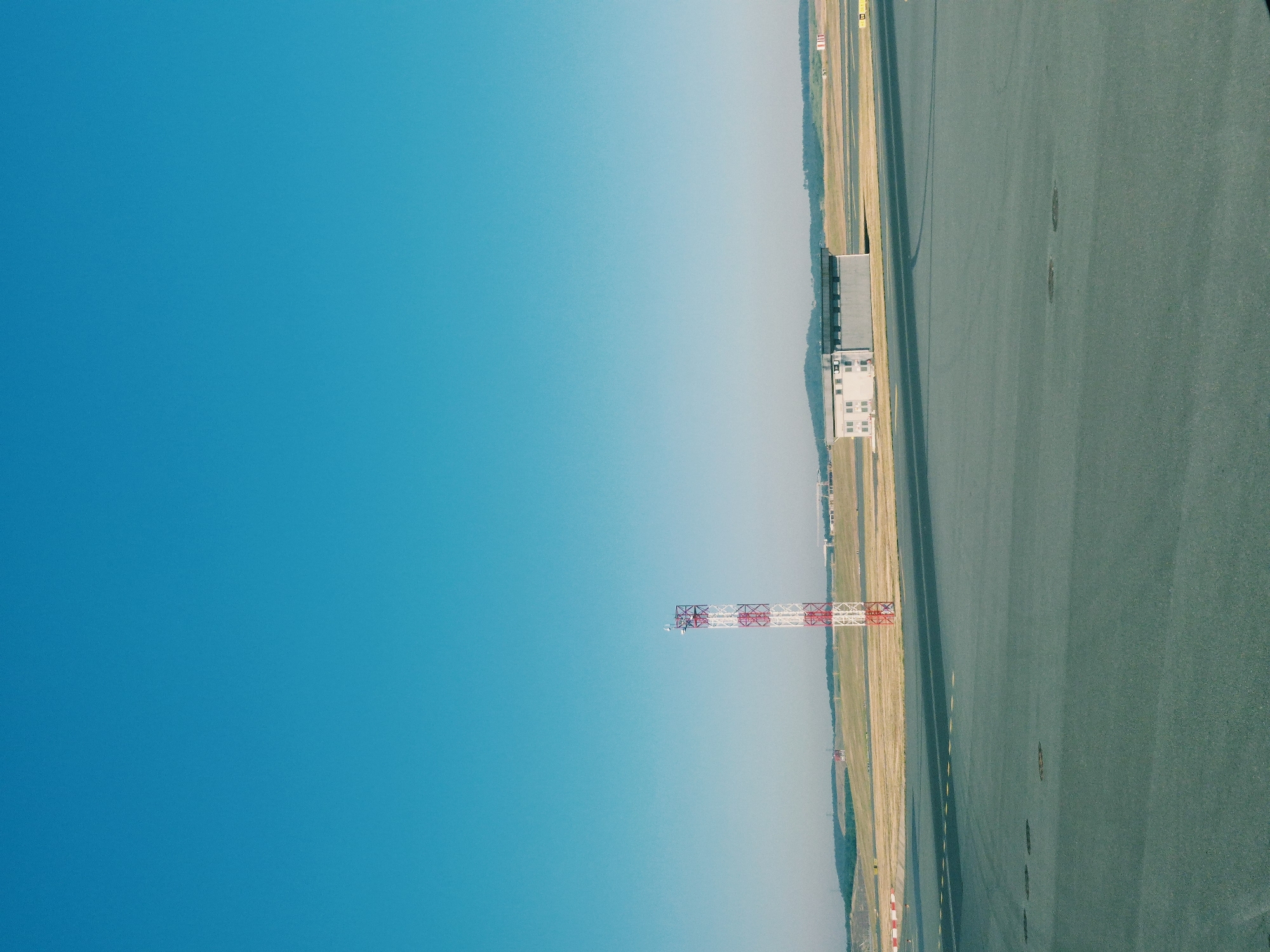 The image depicts an airport tarmac on a clear, sunny day in Arnavutköy, Turkey. A tall, red and white checkered tower, likely an aviation beacon or communication mast, stands prominently on the left side of the frame. In the middle ground, a long, rectangular building with multiple windows and a dark roof is visible, situated amidst flat, grassy terrain. The foreground is dominated by the dark grey asphalt of the runway or taxiway, marked with faint yellow lines. The background shows a gentle, rolling landscape of hills and fields under a vast, bright blue sky. There are no people or vehicles visible in the scene.