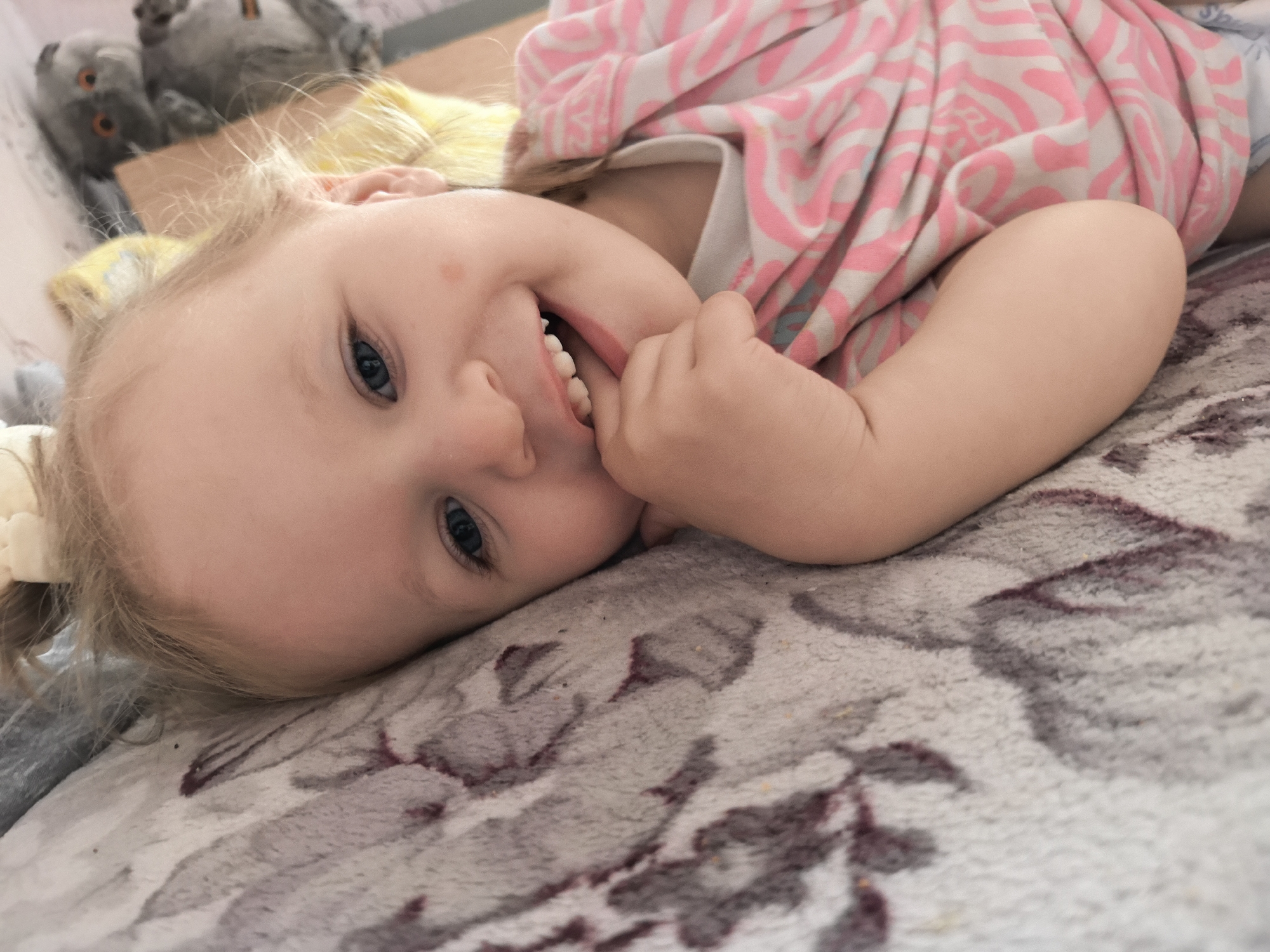 This close-up shot captures a cheerful young child, likely a toddler or preschooler, lying on their side and looking directly into the camera. The child has fair skin, light blonde hair, and bright blue eyes, with a small red mark or freckle visible on their right cheek. They are smiling broadly, showing their teeth, and their left hand is positioned near their mouth, suggesting a playful or curious gesture. The child is wearing a pink and white patterned top or blanket with an abstract or wavy design.
They are resting on a soft, textured surface, which appears to be a patterned rug or blanket featuring shades of grey, white, and maroon or deep purple in an organic, perhaps floral, motif. In the blurred background, two grey owl-shaped stuffed animals or toys with distinct orange eyes are visible, indicating an indoor setting, likely a child's bedroom or play area. The image conveys a joyful and innocent emotion, highlighting the child's happy expression. No visible text is present. While the location context is Chaikovsky, Russia, there are no specific visual cues within the image to confirm this.