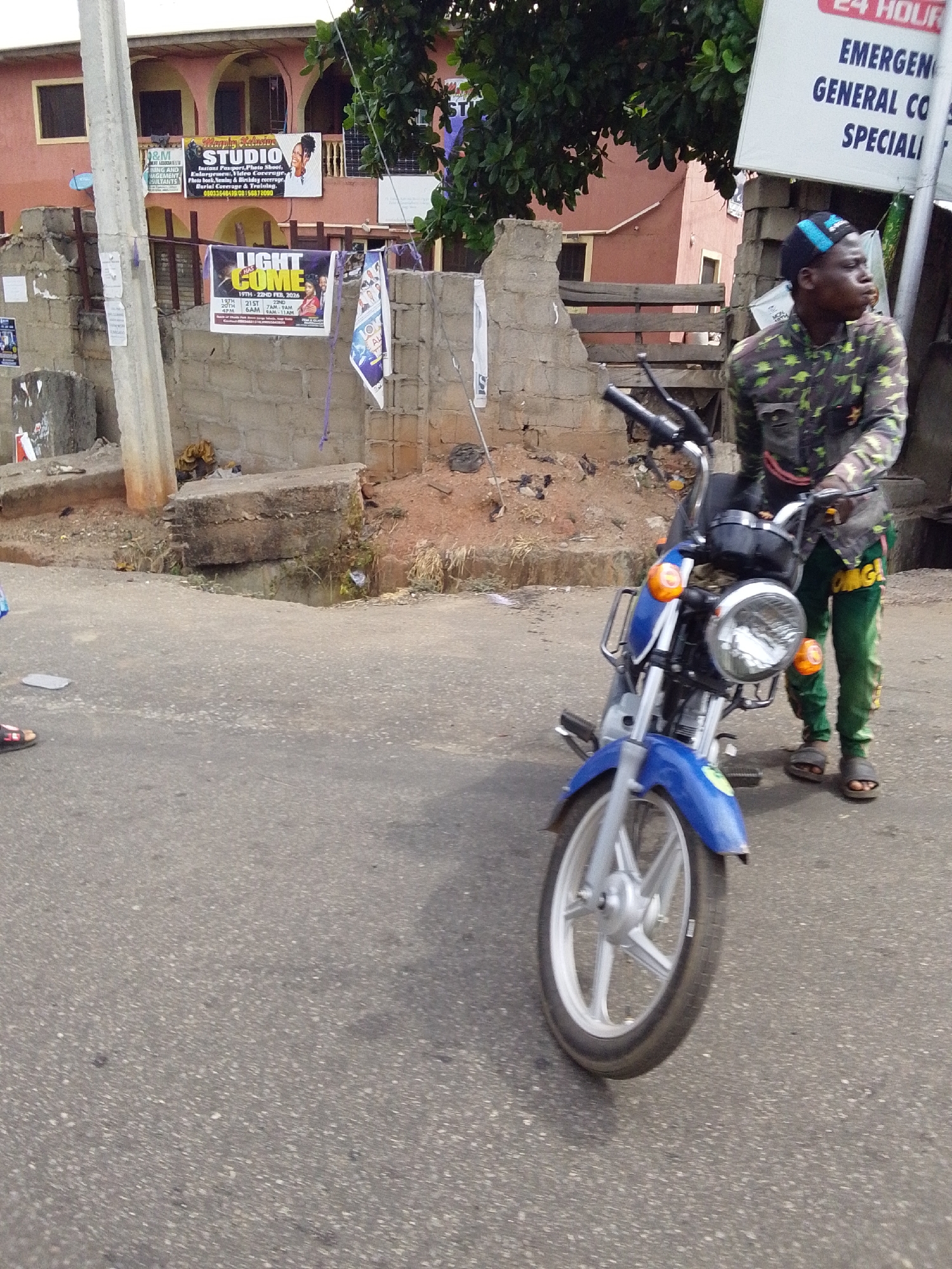 The image, taken in Lokoja, Nigeria, depicts a young man standing next to a blue motorcycle on an asphalt road. He is wearing a camouflage-patterned shirt, green pants with yellow lettering, and sandals. He has a blue cap on his head. Behind him is a partially constructed wall with posters and advertisements, including one for "Murphy Kalusive Studio" and another for "Light Come," dated February 19th-22nd, 2026. A sign advertising a "24 Hour Emergency General" service is visible to his right. A concrete drainage ditch runs alongside the road. The background shows a peach-colored building.