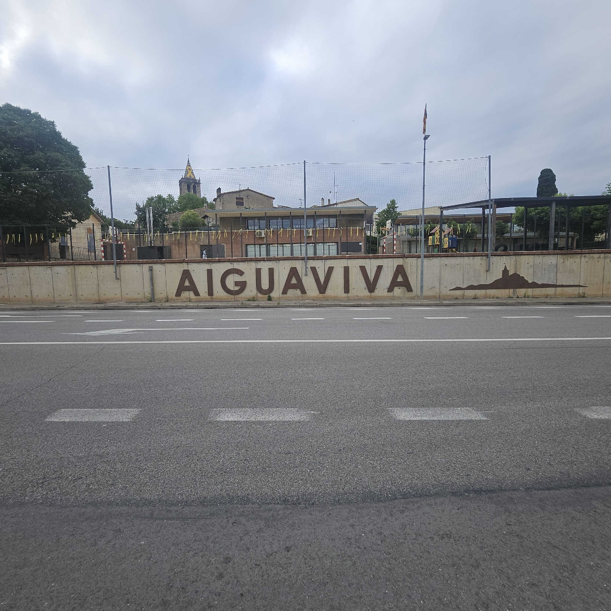 The image shows a view of the town of Aiguaviva, Spain, from across a road. The word "AIGUAVIVA" is prominently displayed in large, dark brown, three-dimensional letters mounted on a concrete wall. To the left of the text, there is a silhouette of a town skyline, likely representing Aiguaviva itself, with a church tower as the most distinct feature. Behind the wall, a building with a flat roof and several windows can be seen, with a sports court or play area visible in front of it, protected by a safety net. A flagpole with a flag is also visible to the right of the building. The foreground consists of a paved road with white lane markings. The sky is overcast with clouds. There are no people clearly visible in the scene.