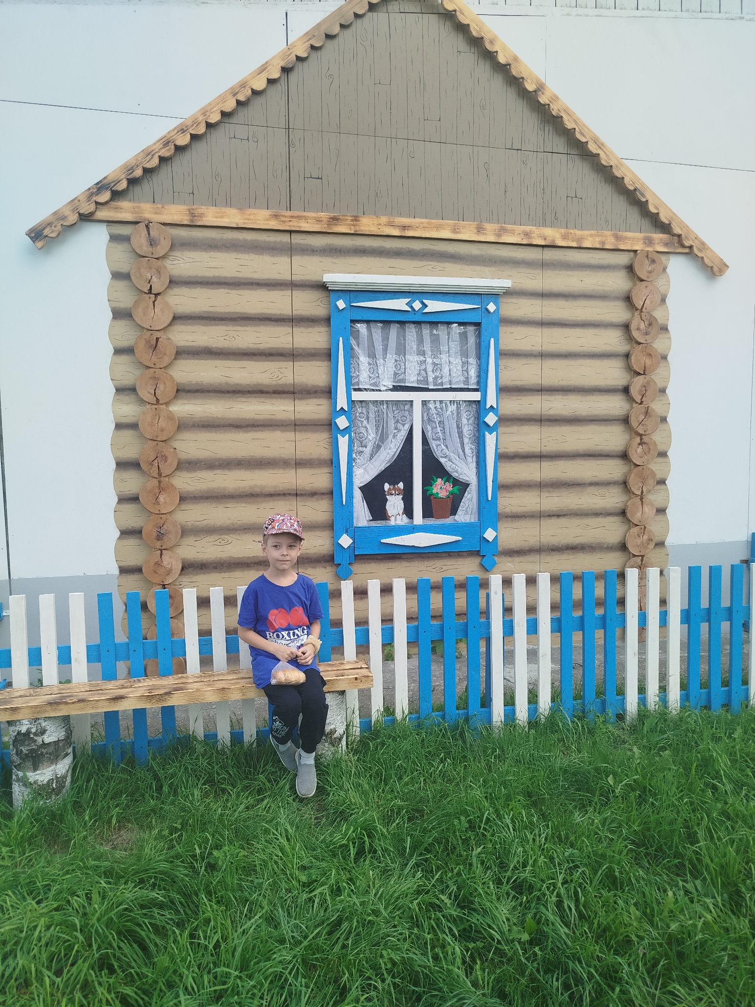 In Zavodoukovsk, Russia, a young boy wearing a blue t-shirt, dark pants, and a patterned cap sits on a wooden bench in front of a mock log cabin facade. He is holding a plastic bag with food inside. The facade features a blue-trimmed window with lace curtains, through which a small cat figure and a potted flower are visible. A decorative fence of white and blue vertical planks separates the bench and the facade from a patch of green grass. The background is a plain wall.