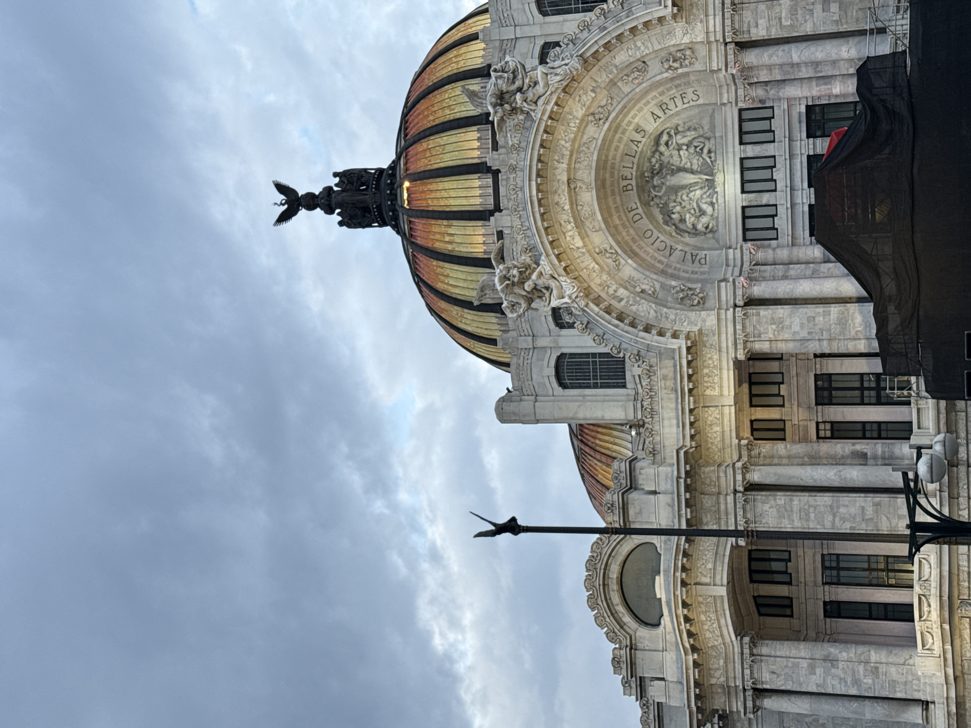 The image depicts a vertical view of the Palacio de Bellas Artes in Mexico City. The building's ornate facade is visible, with its dome featuring gold and copper-colored stripes. Sculptural details adorn the dome's base, and an eagle statue stands at its apex. Part of the building's exterior wall is visible, showcasing intricate carvings and architectural features. The inscription "PALACIO DE BELLAS ARTES" is clearly seen on the building's facade. The sky is overcast with grey clouds. A lamppost with bird-like ornaments on the pole extends into the frame from the right. A black draped fabric partially covers the right side of the building, obstructing some of the windows.
