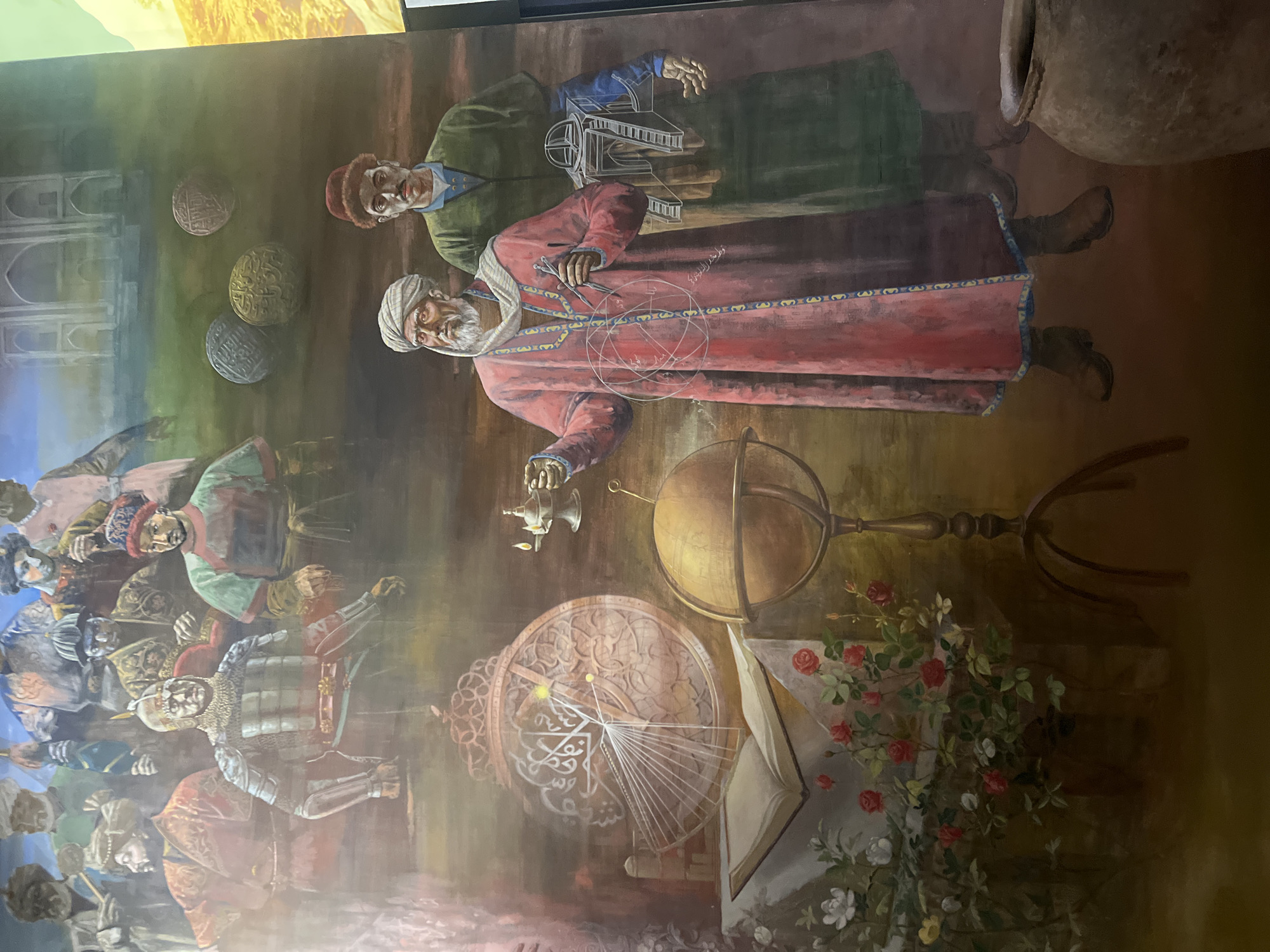 This is a detailed oil painting depicting a historical scene with multiple figures and objects arranged in a complex composition. In the foreground, an elderly man with a white beard and turban stands to the left, wearing a vibrant pink robe adorned with blue and yellow trim. He holds a compass and appears to be engaged in a mathematical or scientific demonstration, with geometric lines superimposed over his attire. To his right stands a younger man in a green tunic and a fur-trimmed hat, holding a blueprint or diagram of an architectural structure.
Behind these central figures, to the left, is a large, ornate astrolabe with intricate markings and a radiating fan-like structure. An open book rests on a table beneath it, surrounded by a cluster of red and white roses. A globe on a stand is positioned between the astrolabe and the men, suggesting themes of astronomy and geography. In the background, a procession of figures in historical attire are depicted, some in armor and other traditional garments, suggesting a gathering or historical reenactment. The backdrop includes architectural elements resembling a grand building, possibly a palace or mosque, and several spheres with intricate carvings are scattered in the middle ground. The overall color palette is rich and earthy, with deep browns, reds, greens, and muted blues dominating the scene. The lighting appears to be soft and diffused, contributing to the painterly and slightly atmospheric quality of the artwork. The painting is likely a mural or a large-scale canvas, given its detailed depiction and the grandeur of the figures and setting. The location is not identifiable.